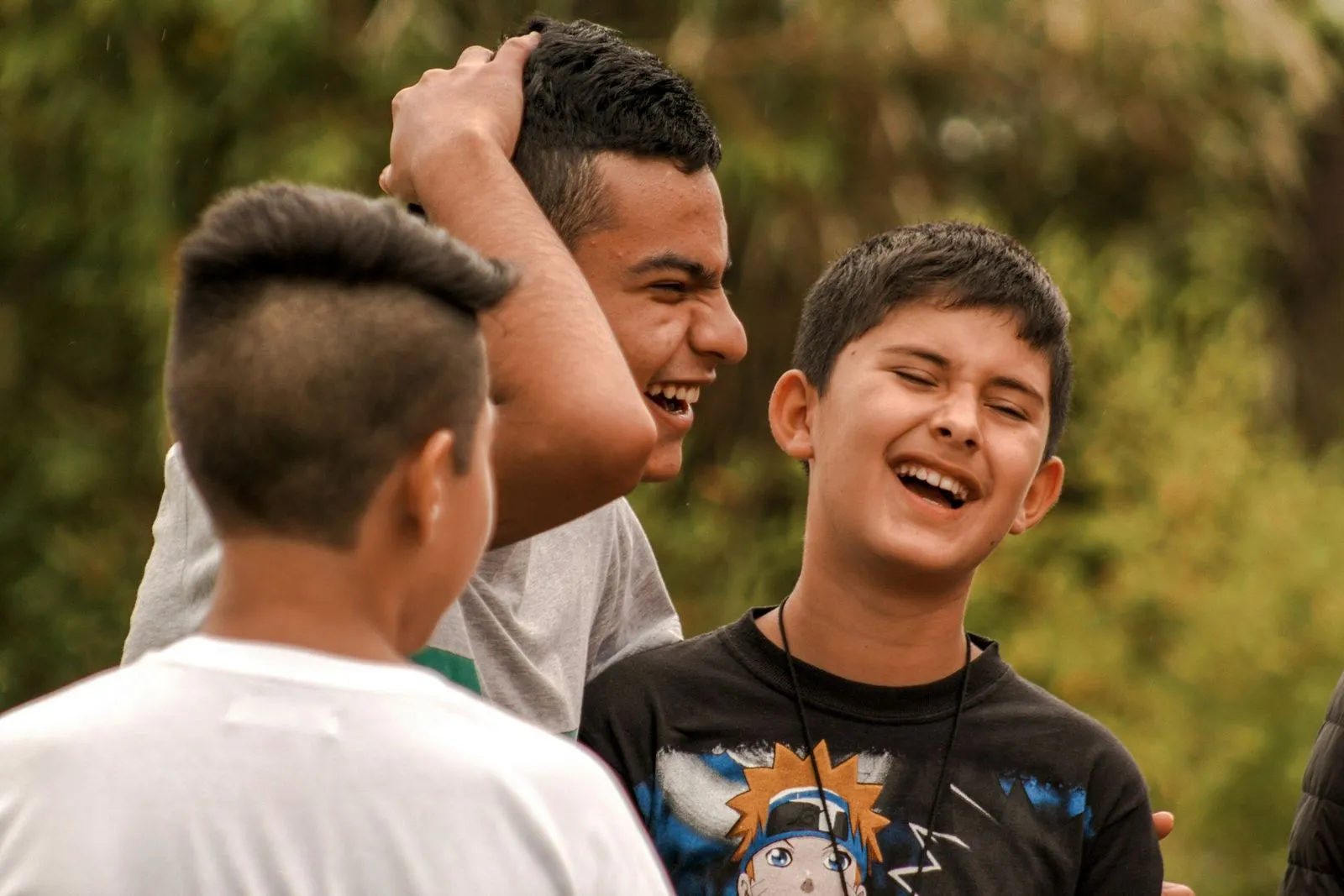 Three boys outdoors, smiling and laughing together. One boy playfully touches another's head. Their joyful expressions convey happiness and friendship.
