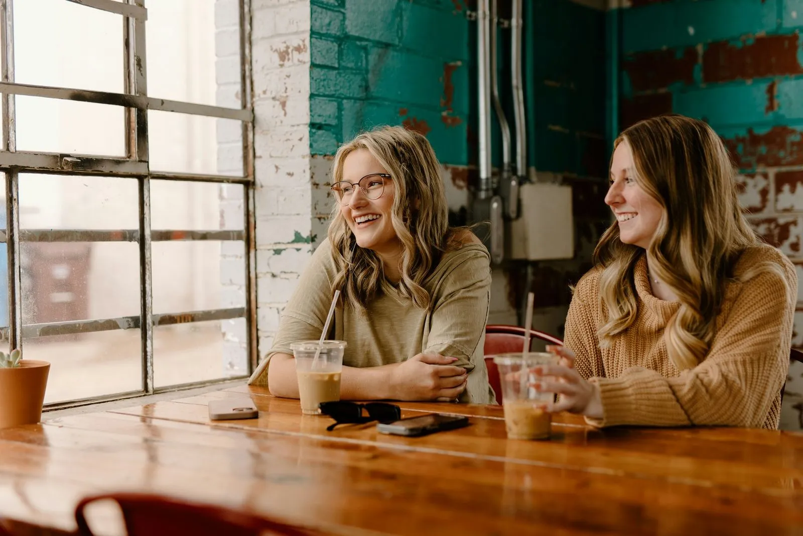Two women sit in a cozy cafe with brick walls, laughing and drinking iced coffee. Warm tones and natural light create a welcoming atmosphere.