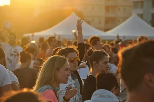 A vibrant outdoor festival at sunset with people gathered in front of white tents. Warm sunlight creates a lively and energetic atmosphere.