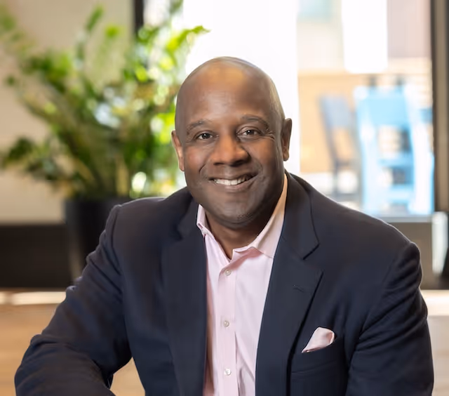Smiling bald man in a dark blazer and light pink shirt seated indoors with blurred green plant and furniture in the background.