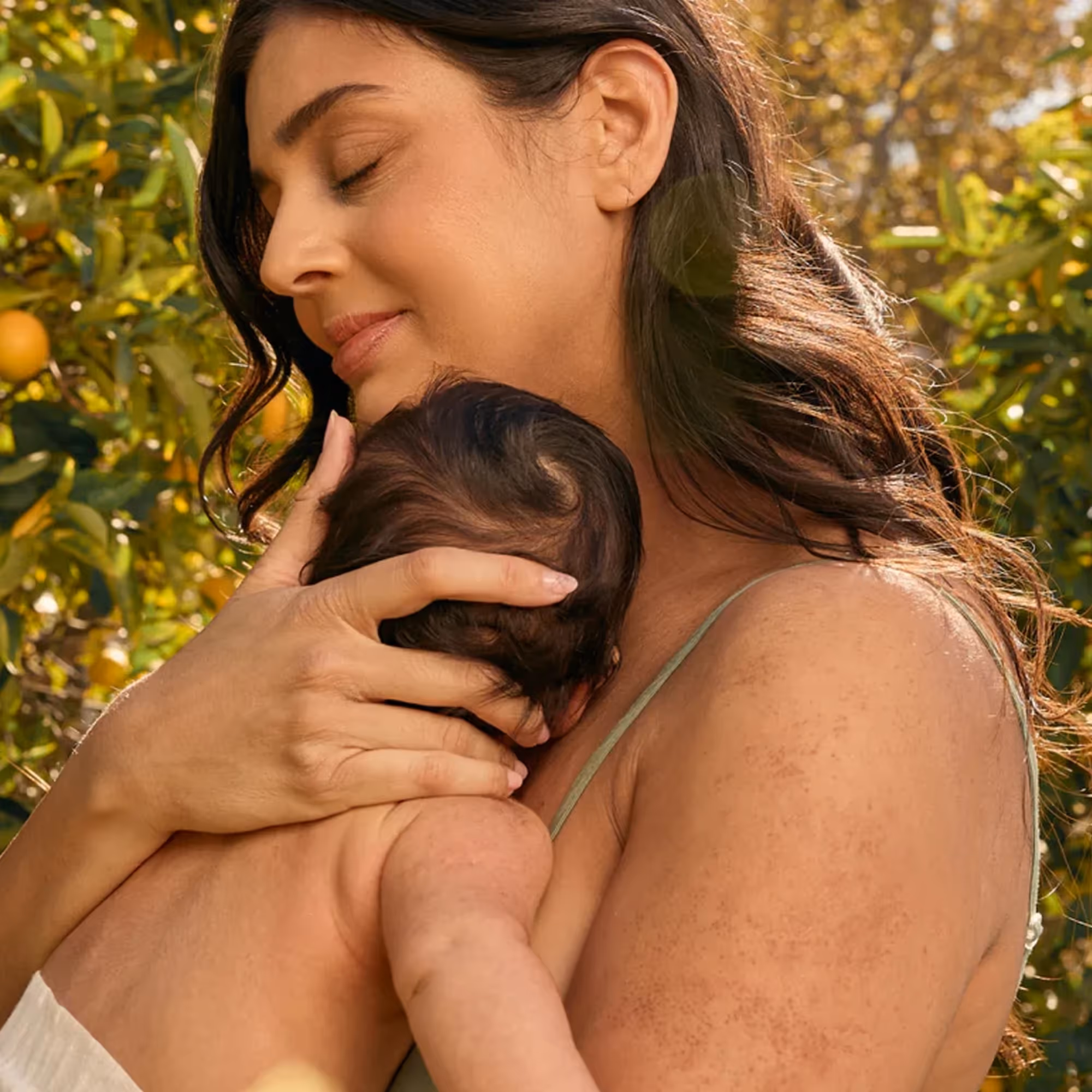 Woman with closed eyes gently holding a newborn baby against her shoulder outdoors with greenery in the background.