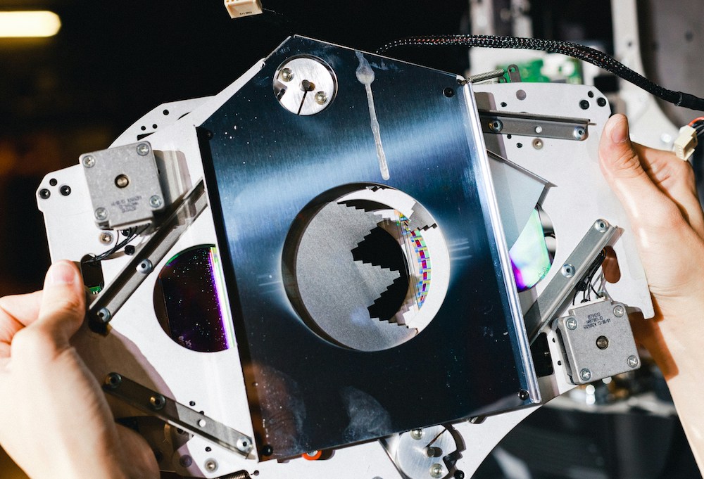 Hands holding a complex mechanical device with mirrored metal plates and visible circuit components.