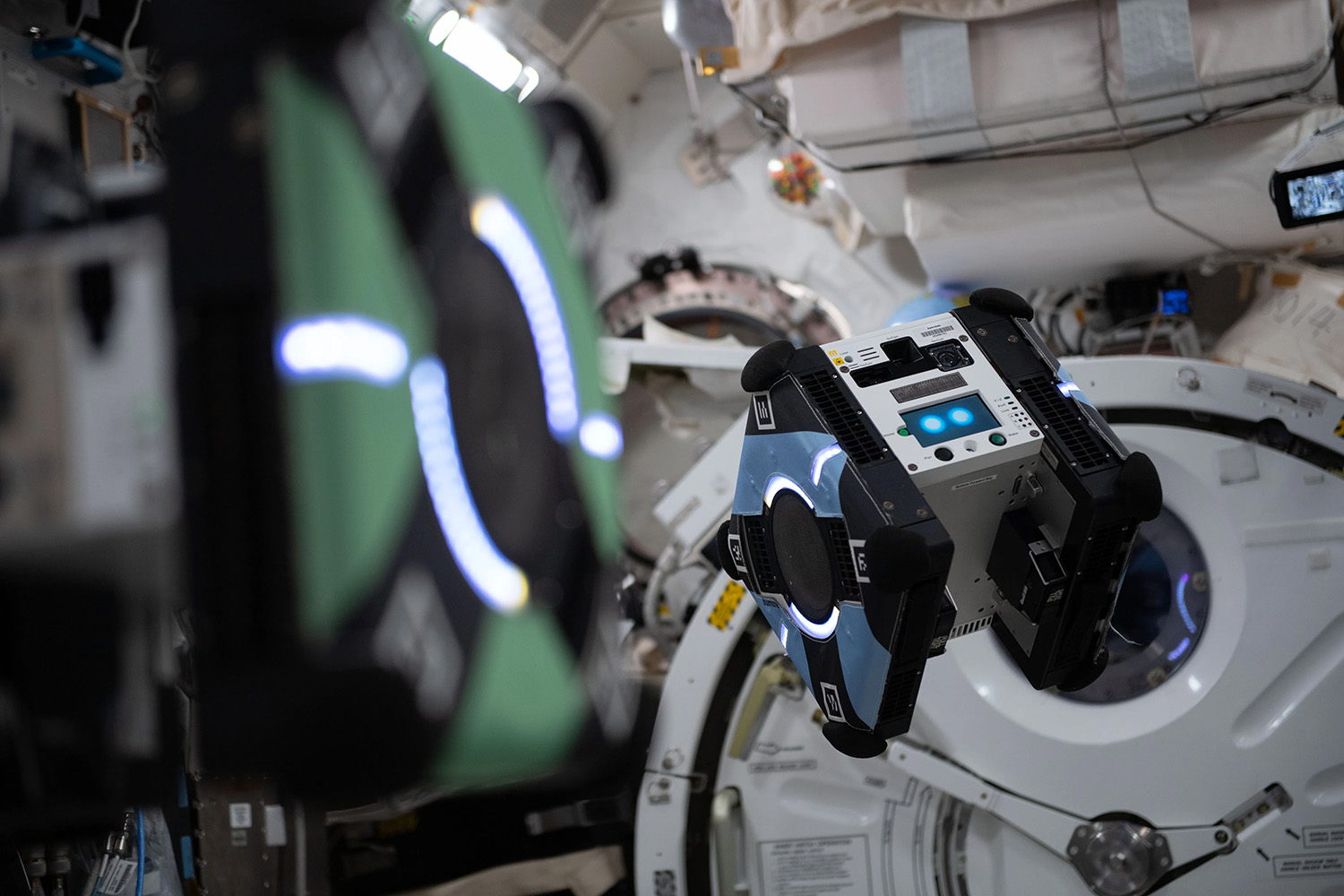 Astrobee robot floating inside a space station module with scientific equipment and storage bags in the background.