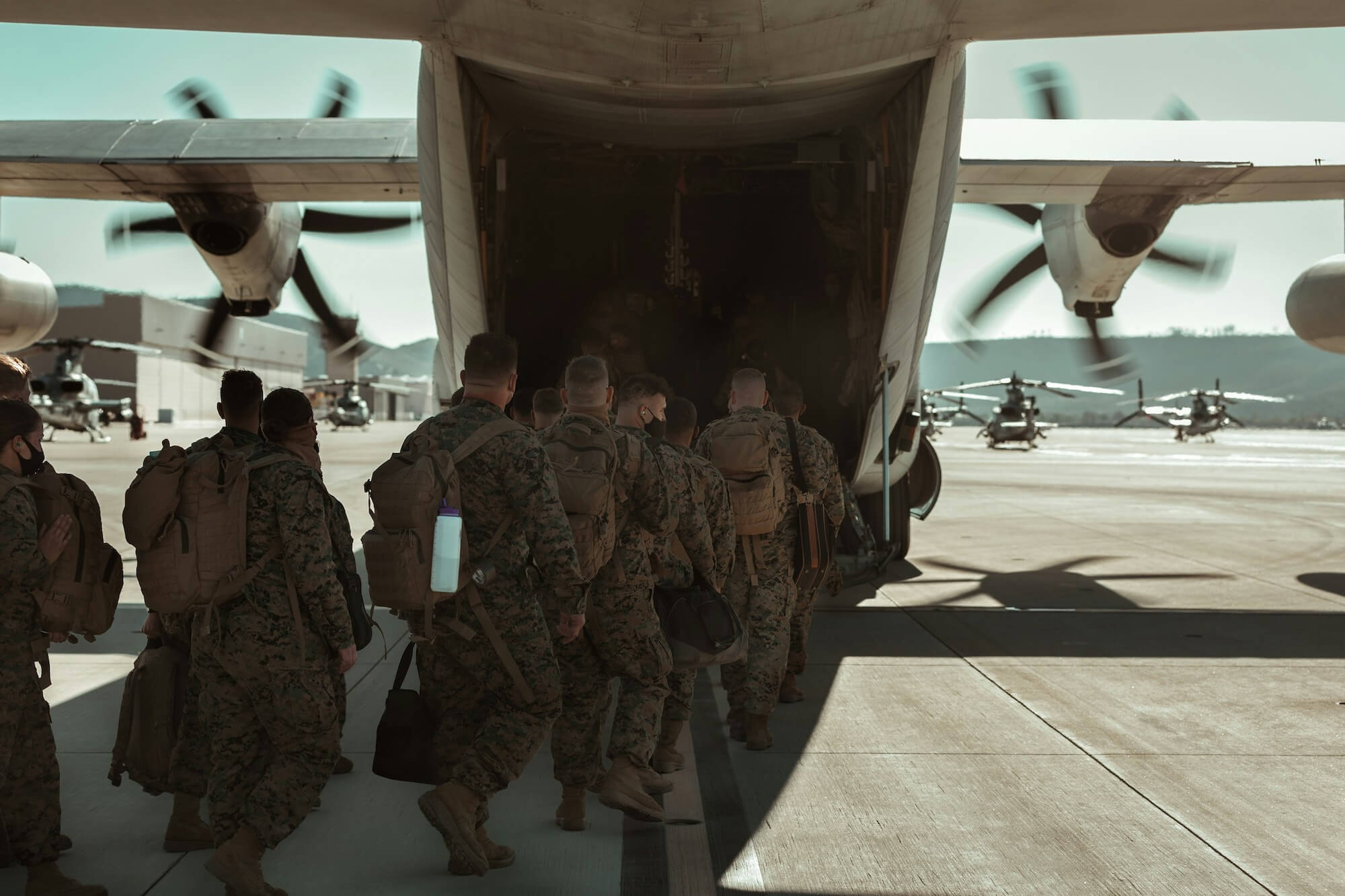 Soldiers in camouflage uniforms and backpacks boarding a military transport aircraft on an airfield with helicopters in the background.