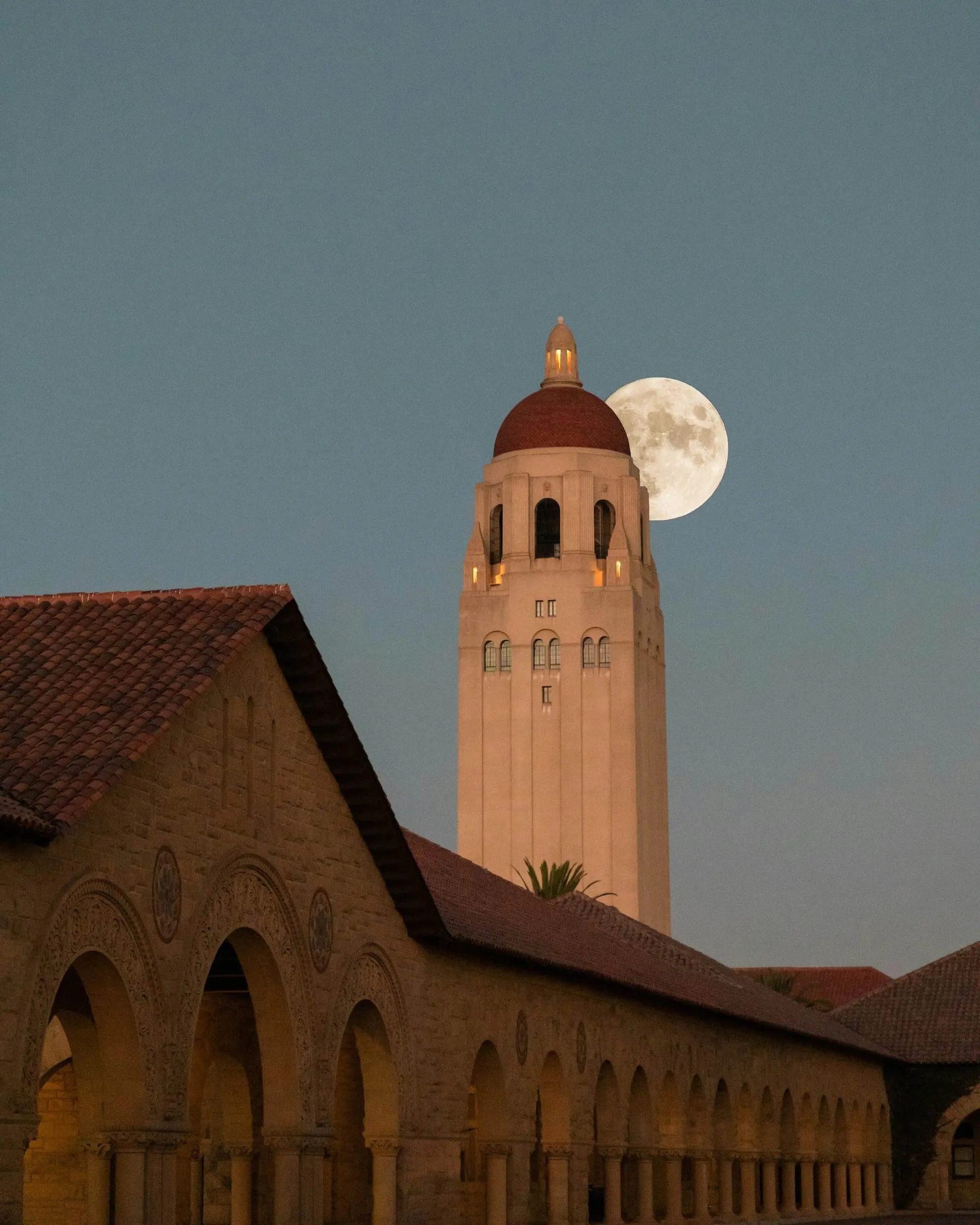 Full moon rising behind the illuminated Stanford University tower at dusk with arched colonnade in the foreground.