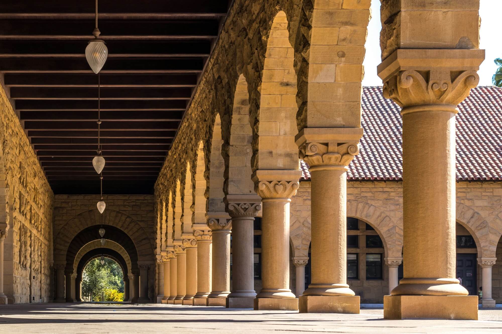 Stone colonnade at Stanford University with arched openings and decorative columns under a wooden-beamed ceiling with hanging lights.