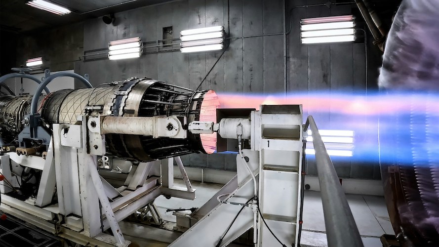 Industrial engine on a test stand emitting a blue and pink flame in a lit testing facility.