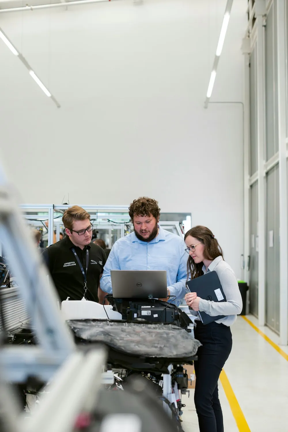 Three engineers, two men and one woman, discussing work while looking at a laptop in a bright industrial lab.