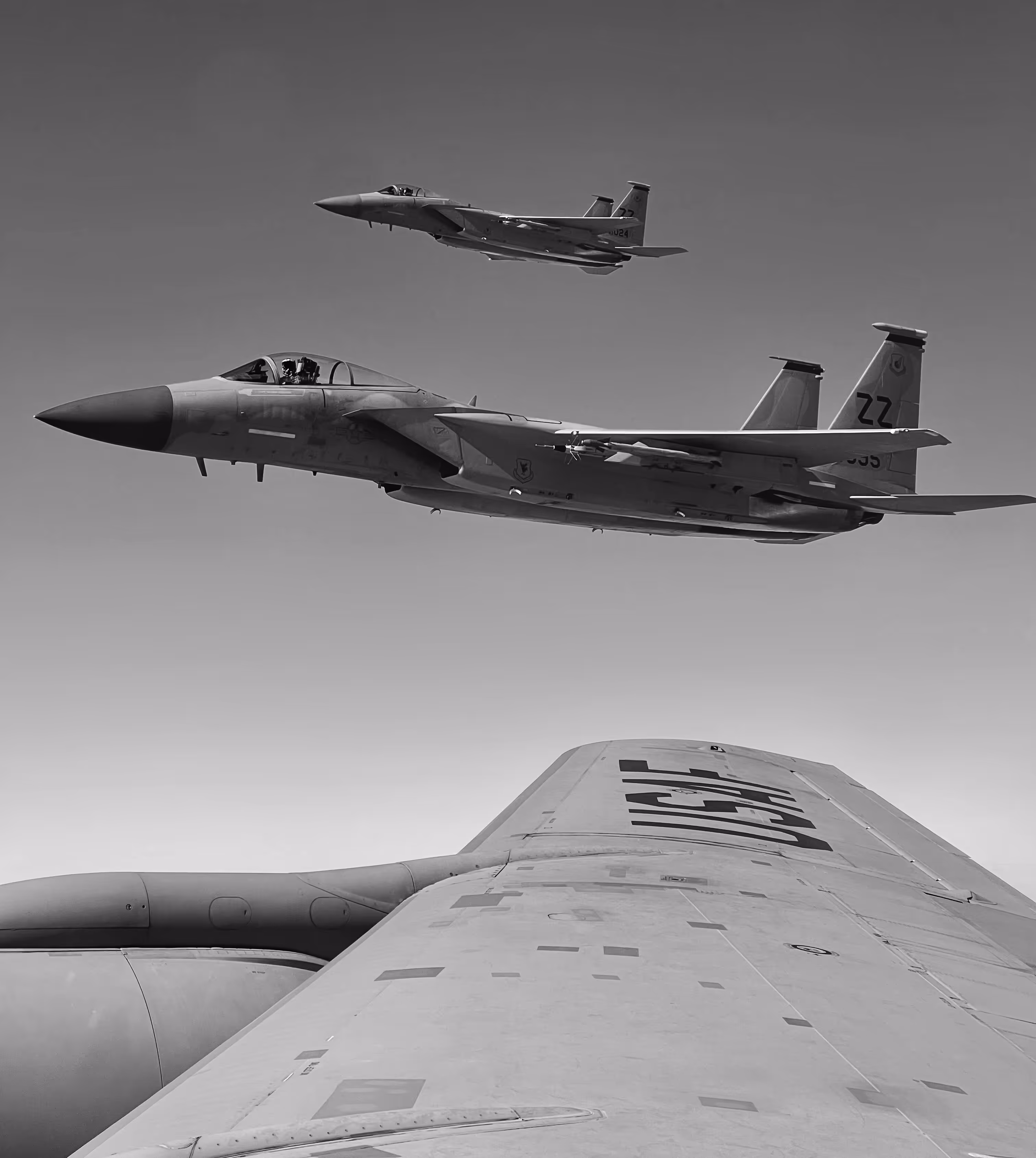 Two F-15 fighter jets flying in formation above the wing of another aircraft.