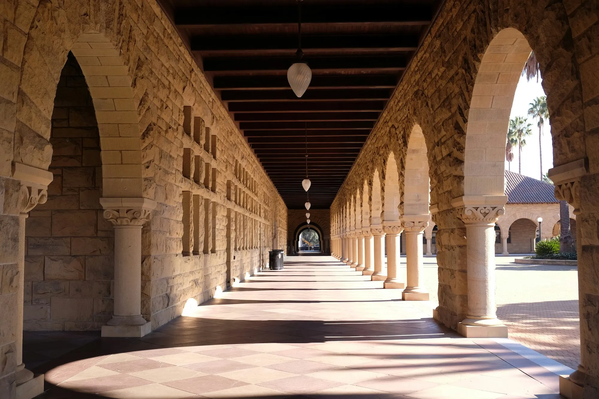 Sunlit stone colonnade with arches, columns, and hanging lamps casting shadows on patterned floor.