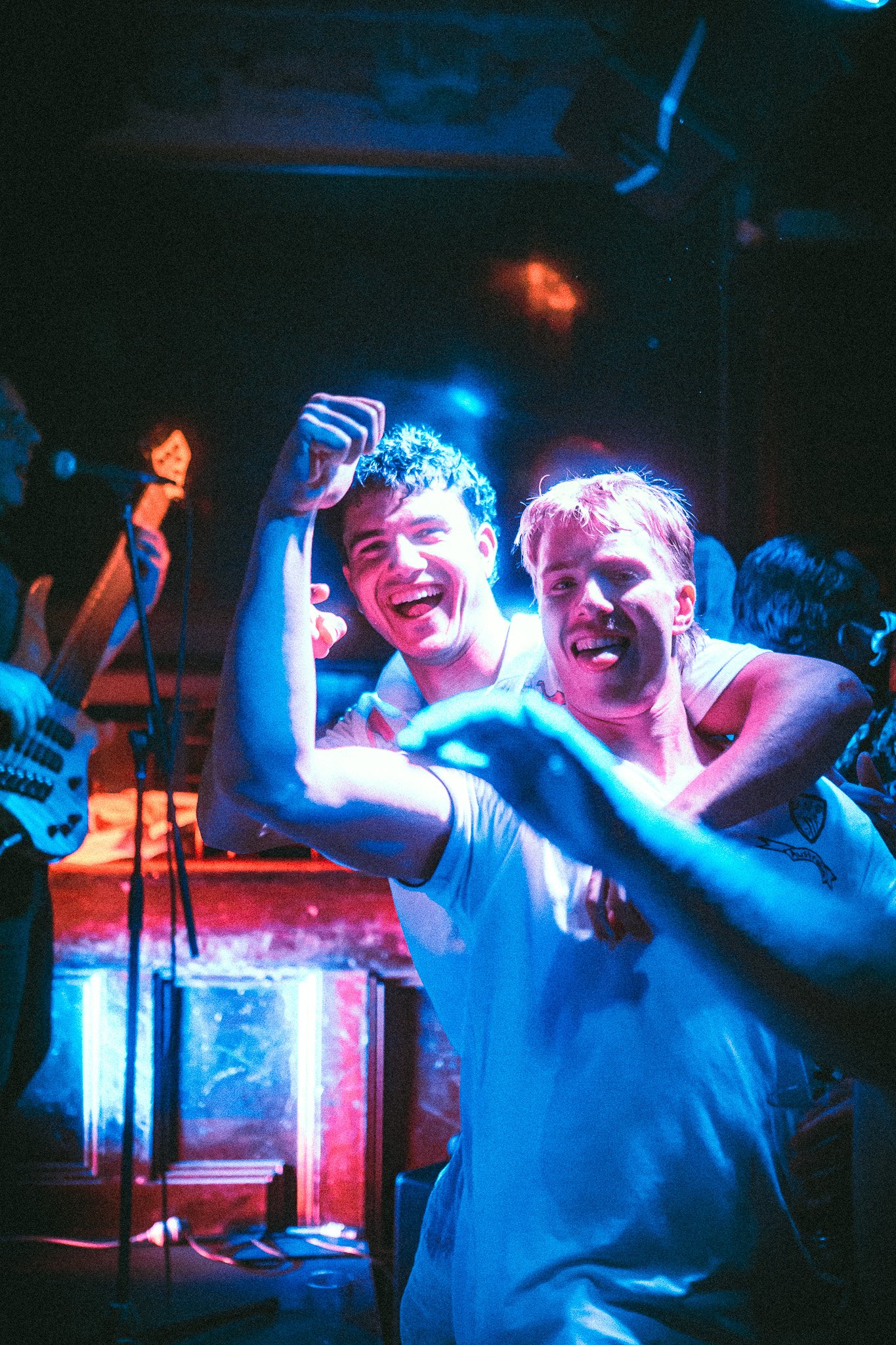 Two young men smiling and posing with flexed arms at a lively music venue with blue and red lighting. LUXE 