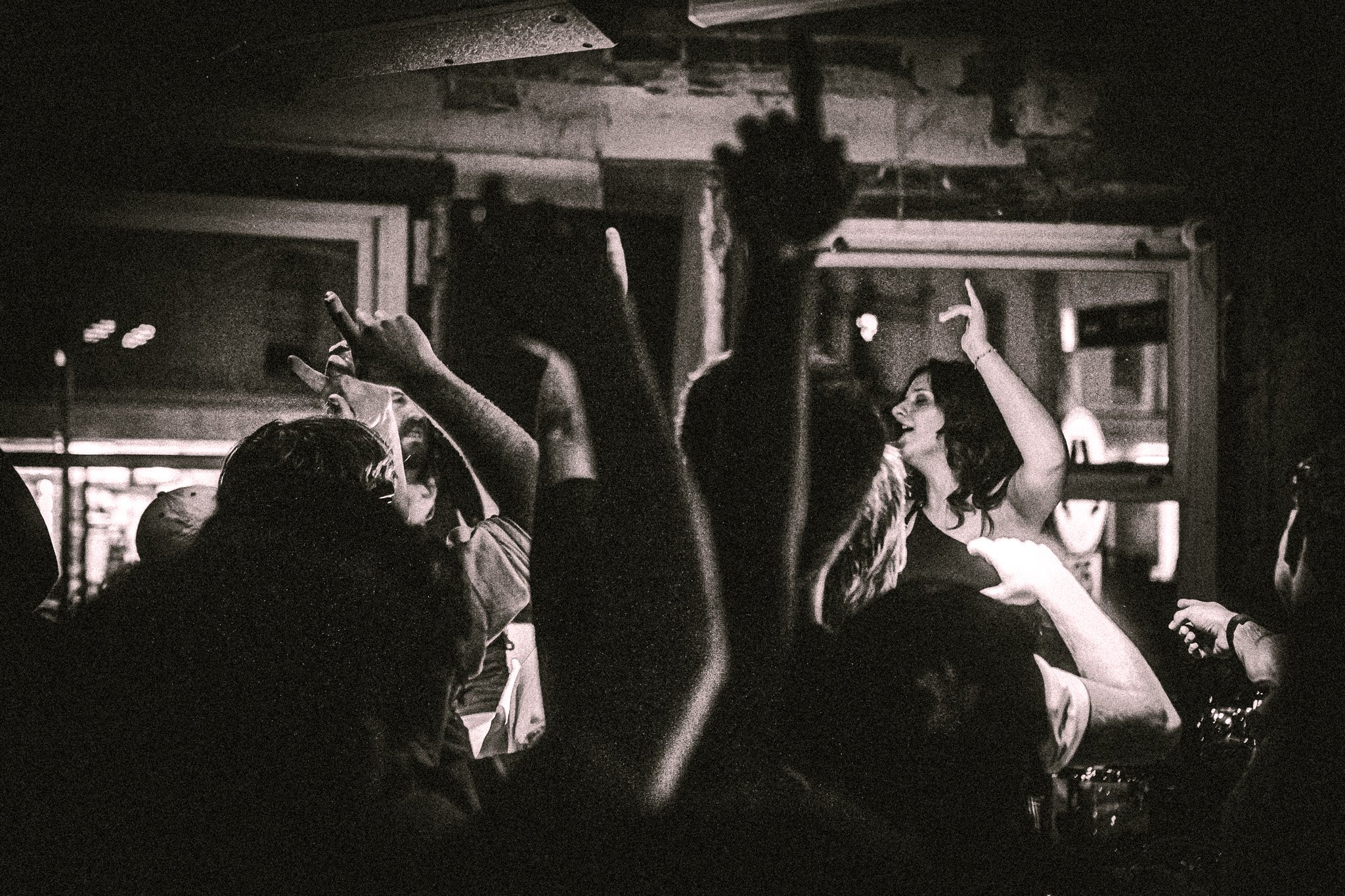 Black-and-white photo of a lively crowd dancing and raising their hands indoors. LUXE 