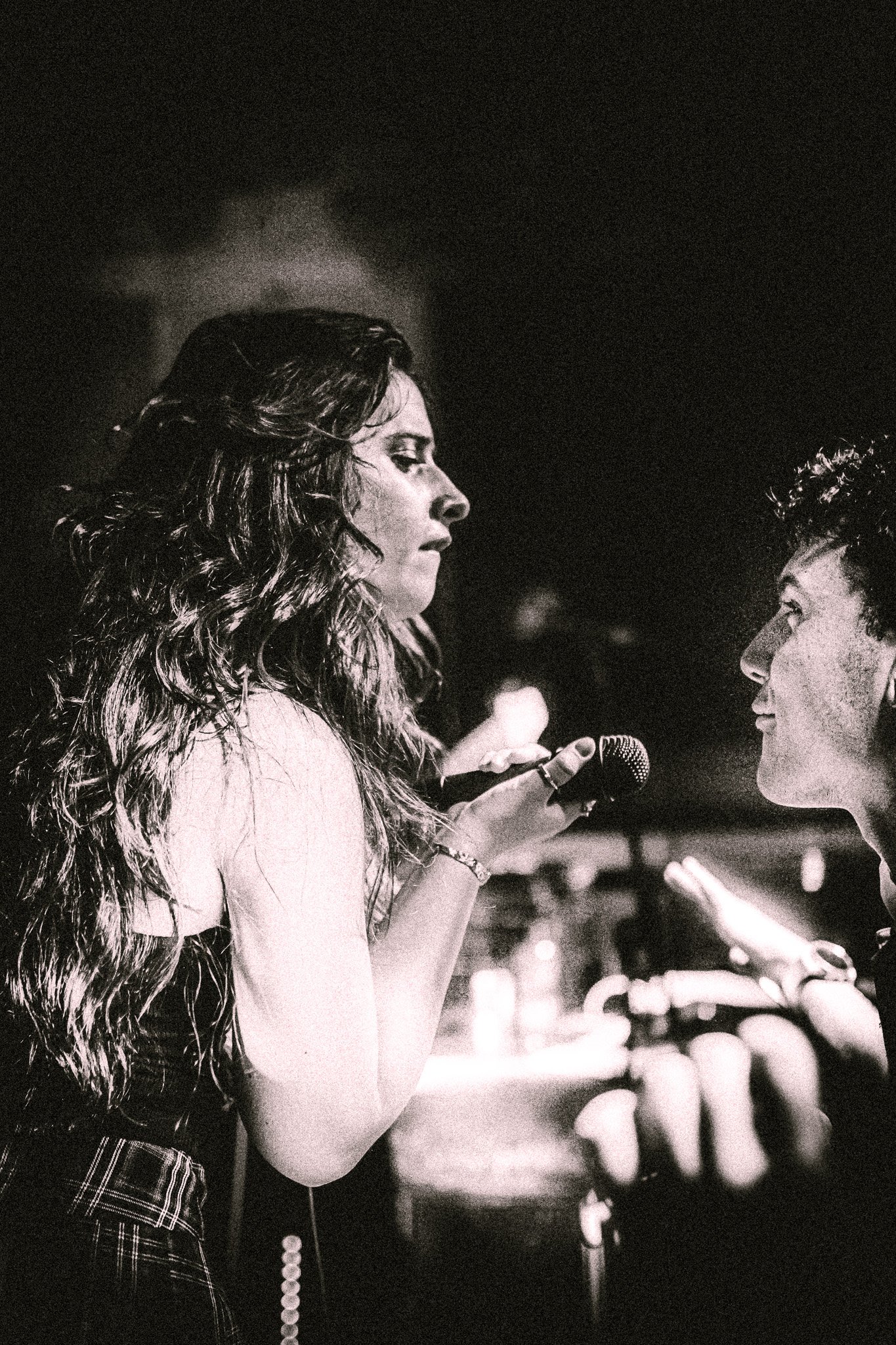 Black and white photo of a woman holding a microphone close to a young man's face in a dimly lit setting. LUXE 