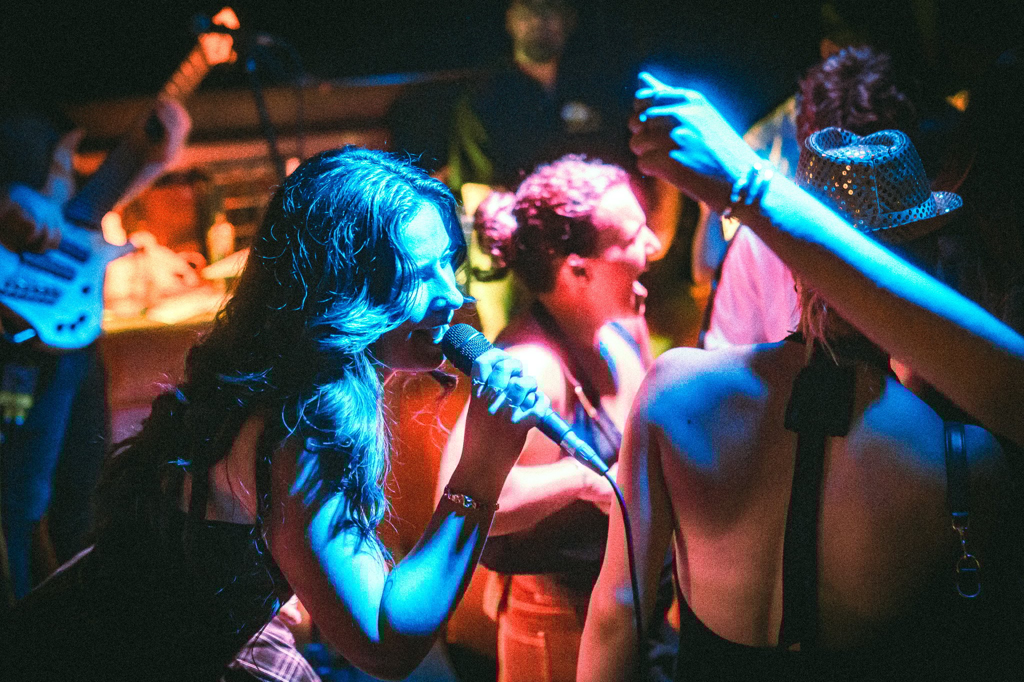 LUXE Woman singing into a microphone on a vibrant stage with other people dancing and a bass guitar player in the background.