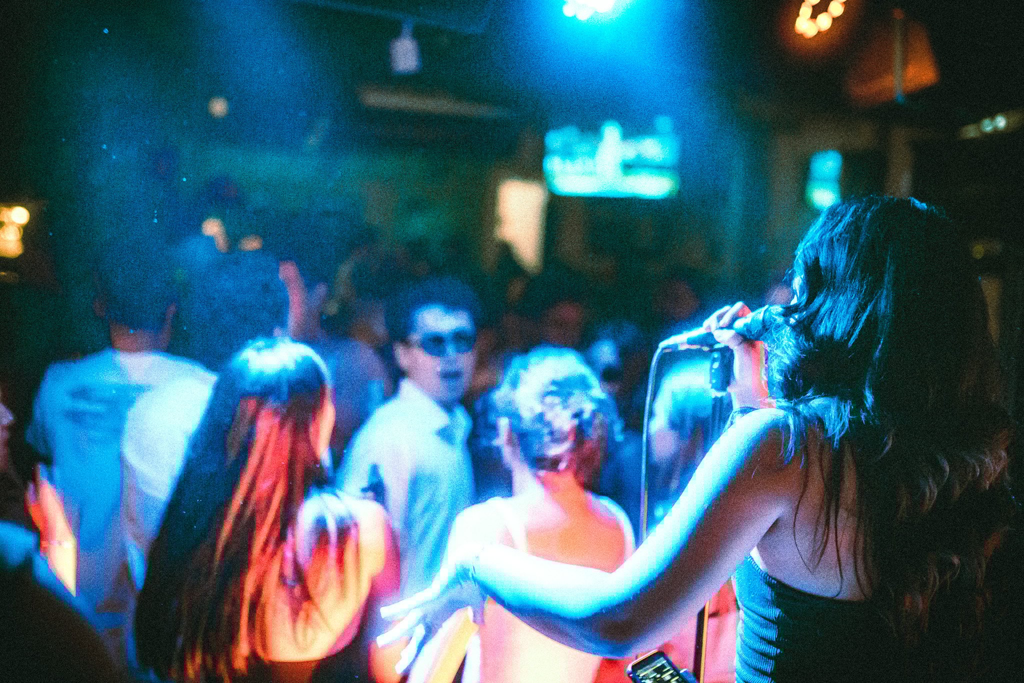 Woman singing into a microphone on stage with colorful lighting and an audience watching. LUXE 