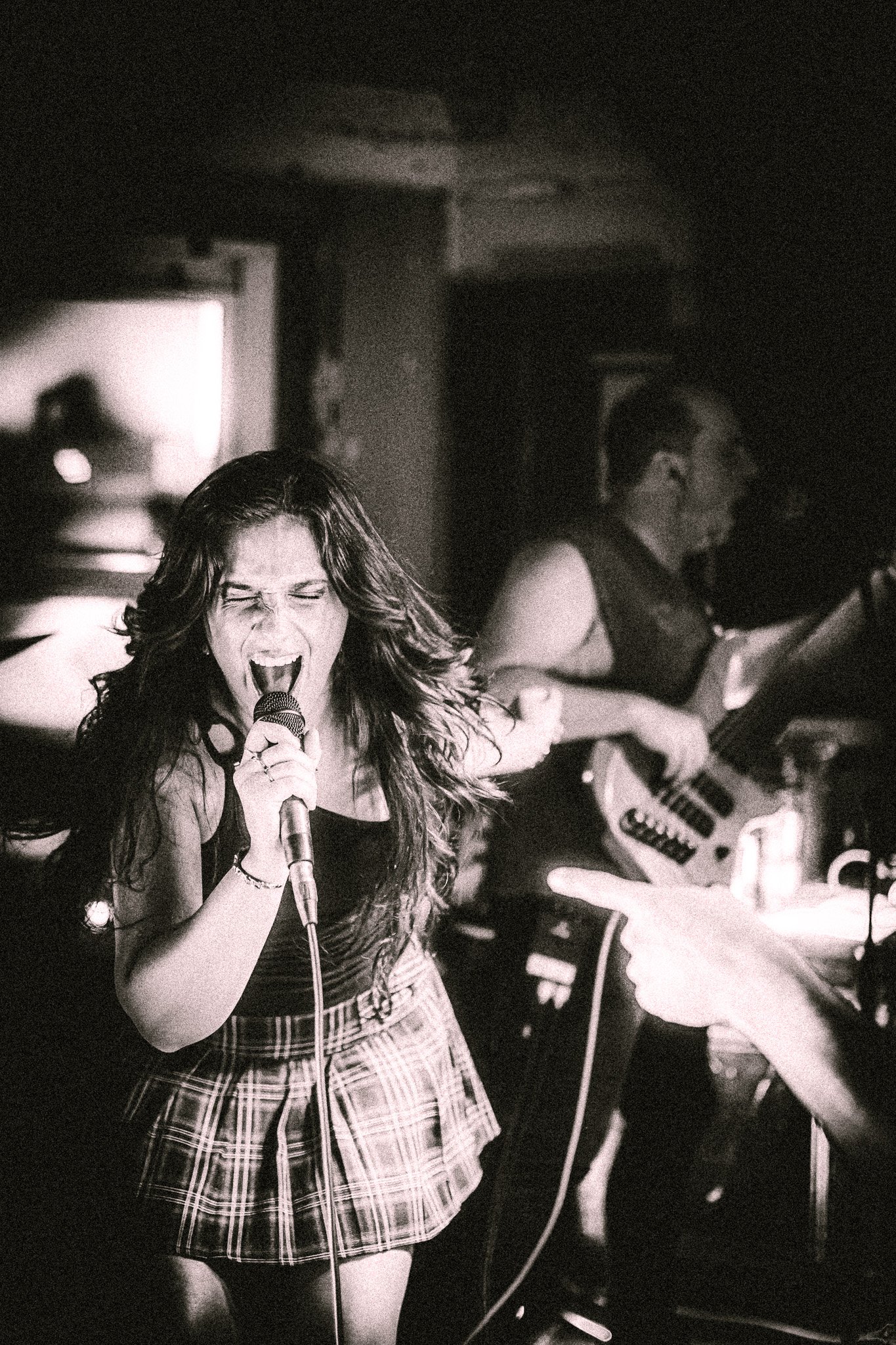 Energetic female vocalist passionately singing into a microphone on stage with a bassist playing in the background. LUXE 