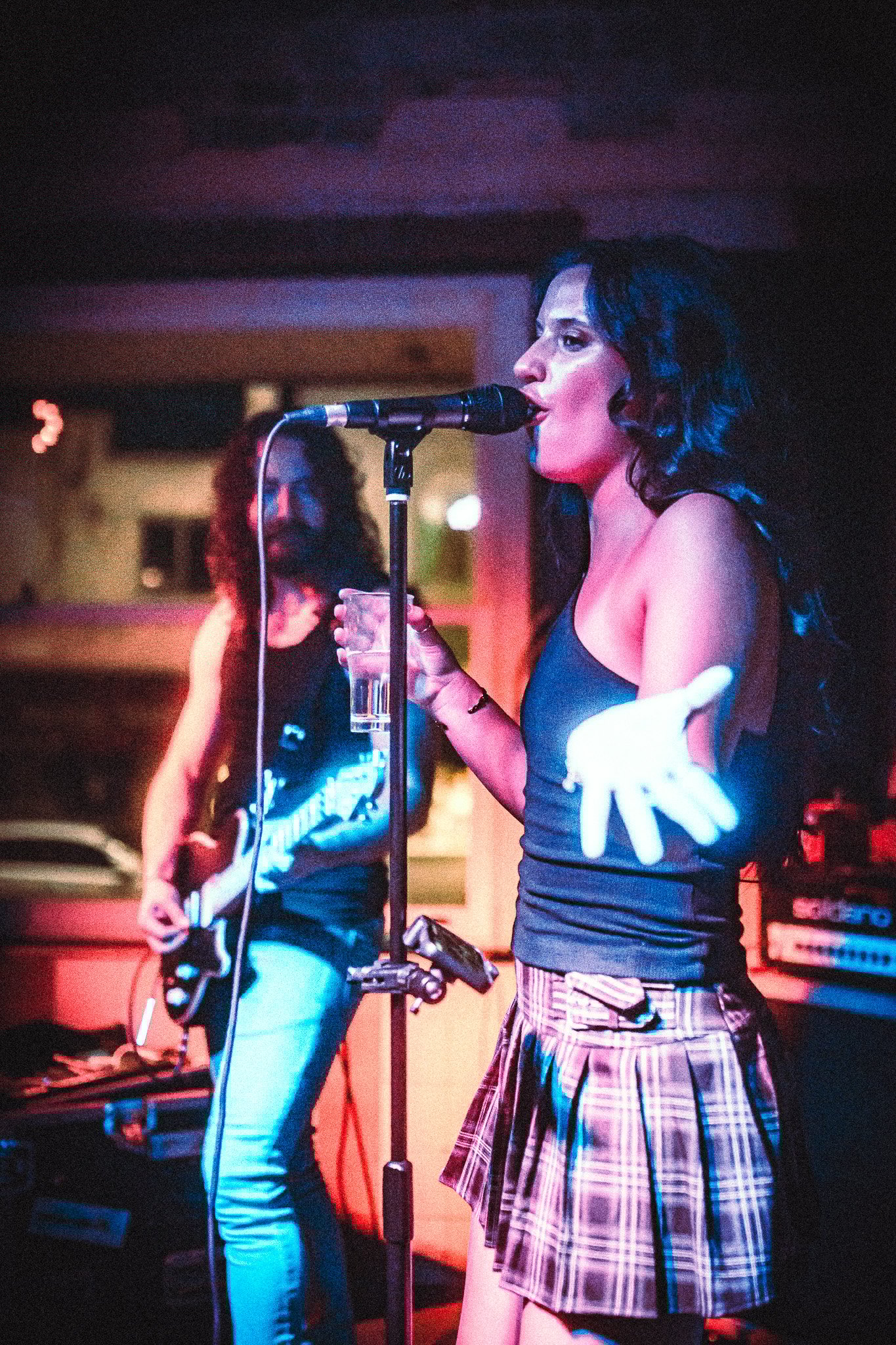LUXE Female singer in a black tank top and plaid skirt passionately performing with a microphone, holding a glass, and a guitarist playing in the background.