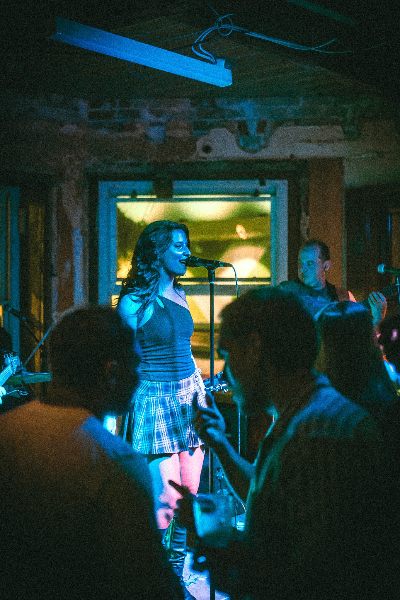 Female singer in a plaid skirt and black top performing on stage with microphone and band in a dimly lit venue. LUXE 