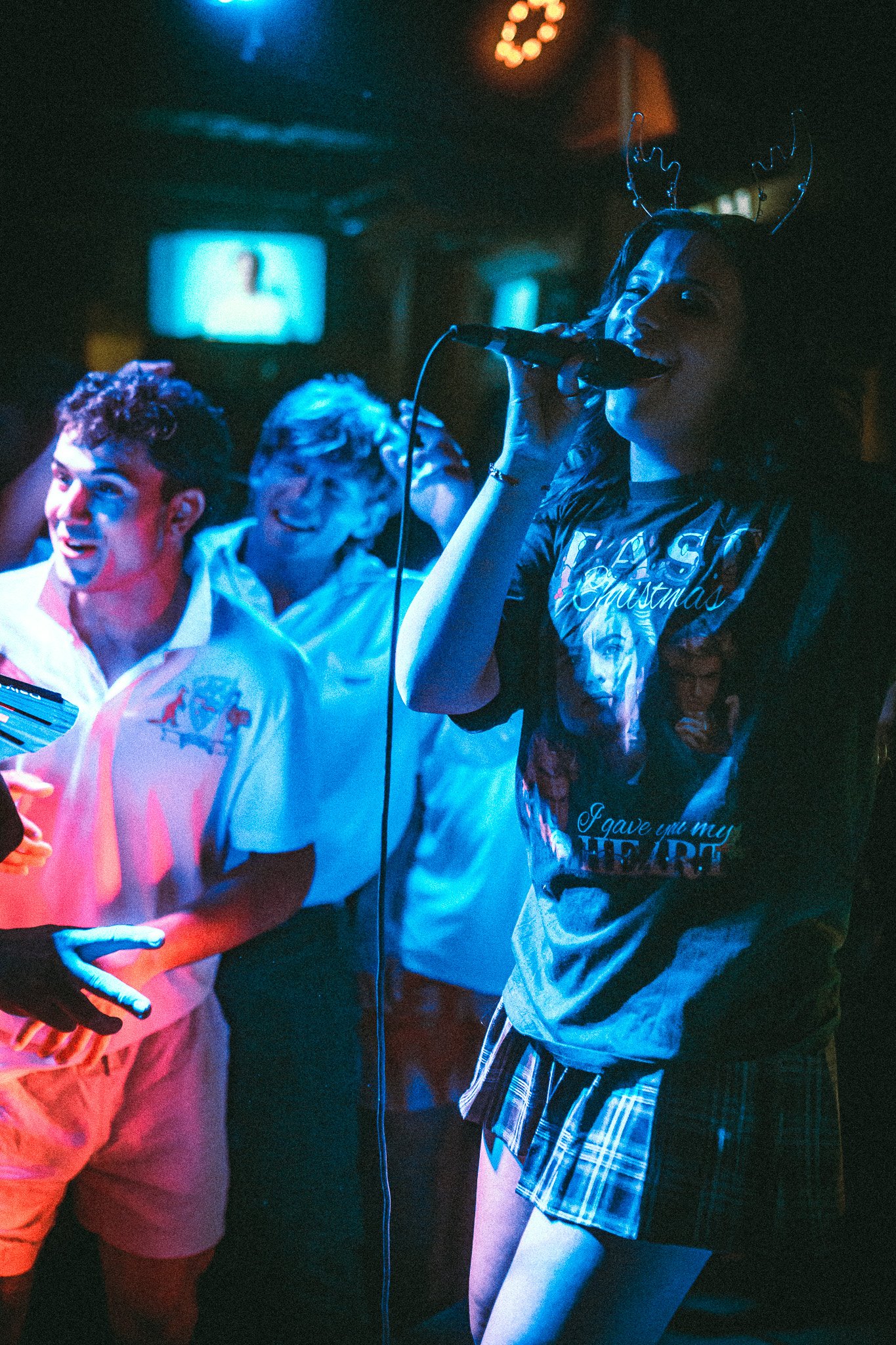 Young woman wearing reindeer antlers singing into a microphone at a lively party with people dancing nearby under colorful lights. LUXE 
