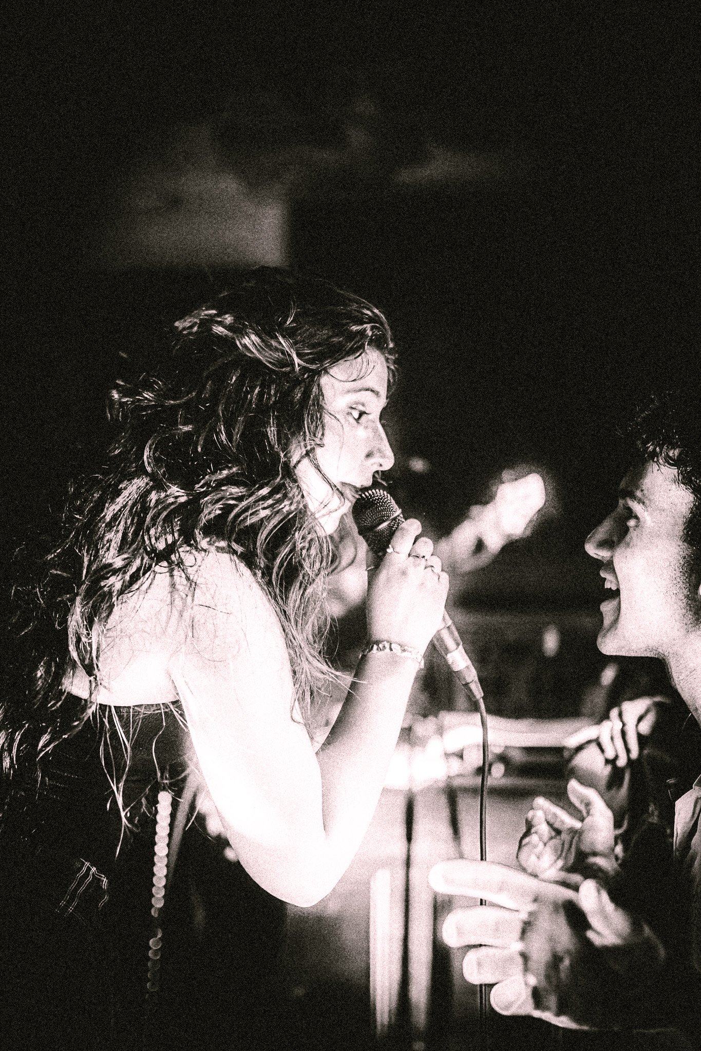 Black and white photo of a female singer with long wavy hair holding a microphone and engaging with an enthusiastic male fan. LUXE 