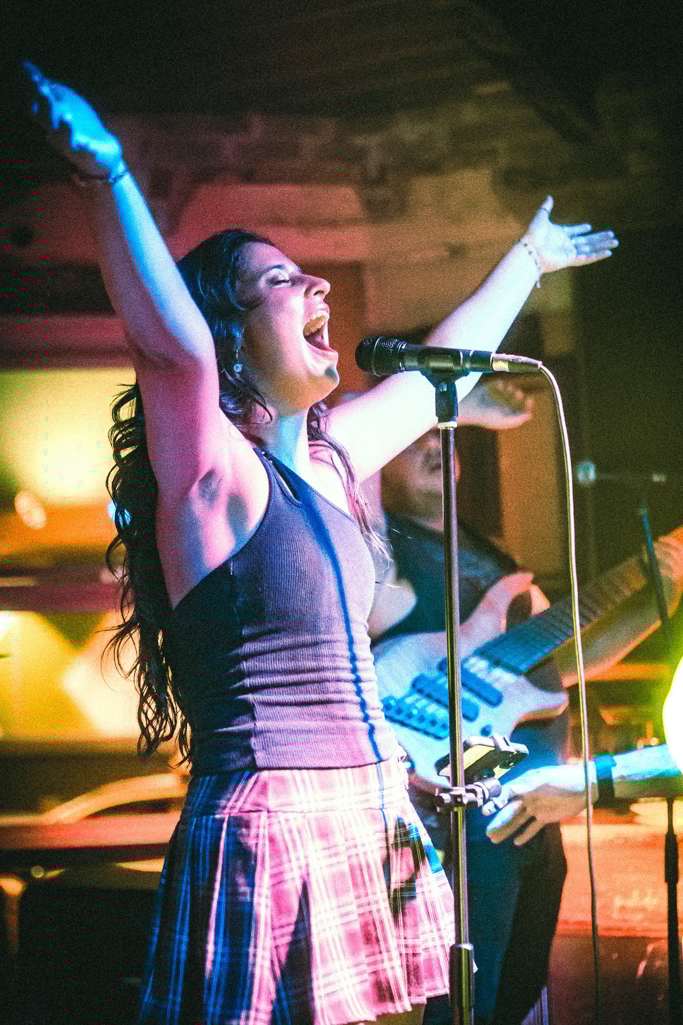 Woman passionately singing with arms raised on stage in front of a microphone, with a guitarist in the background. LUXE 