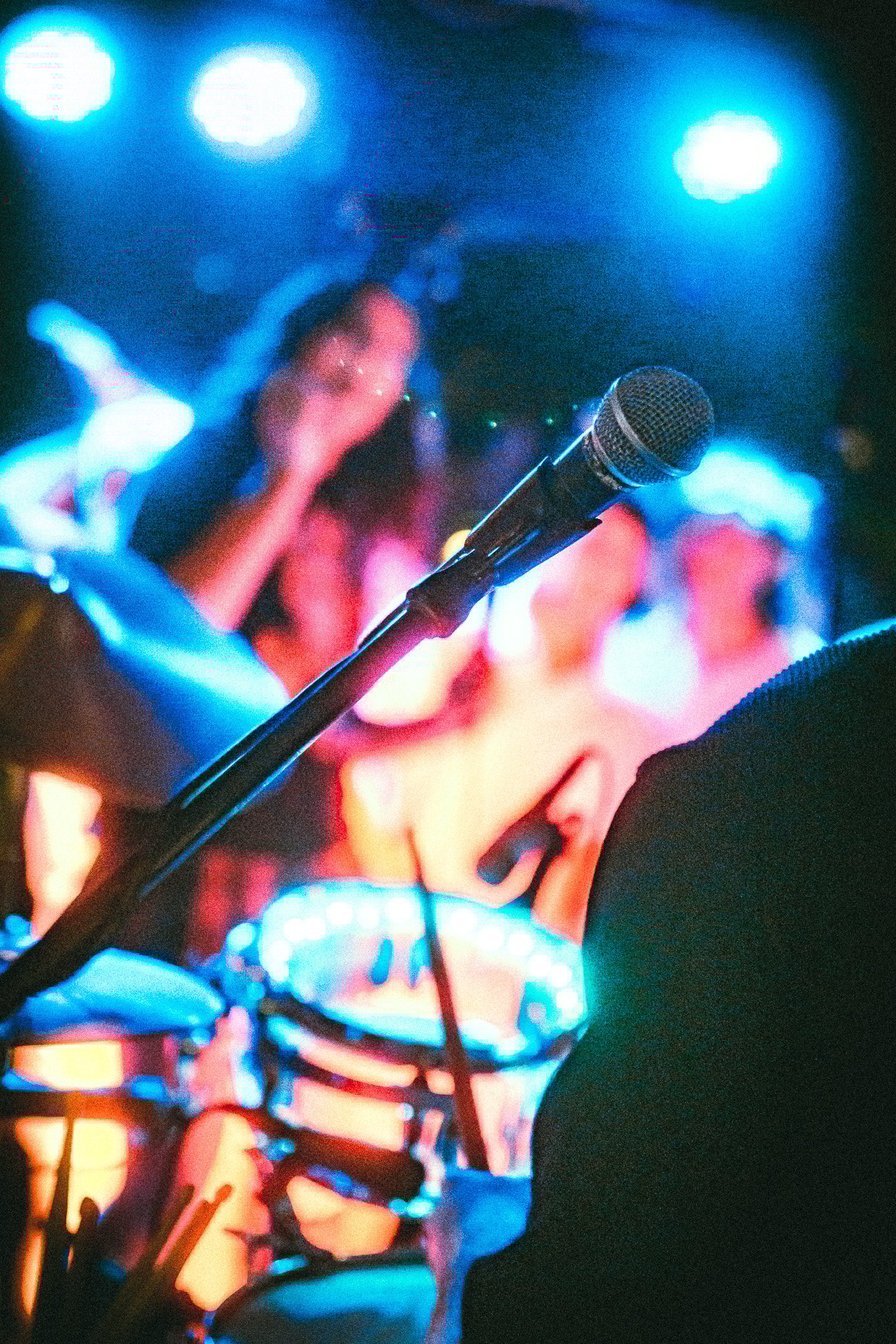 Close-up of a microphone on stage with colorful blurred musicians and drums in the background under blue stage lights. LUXE 