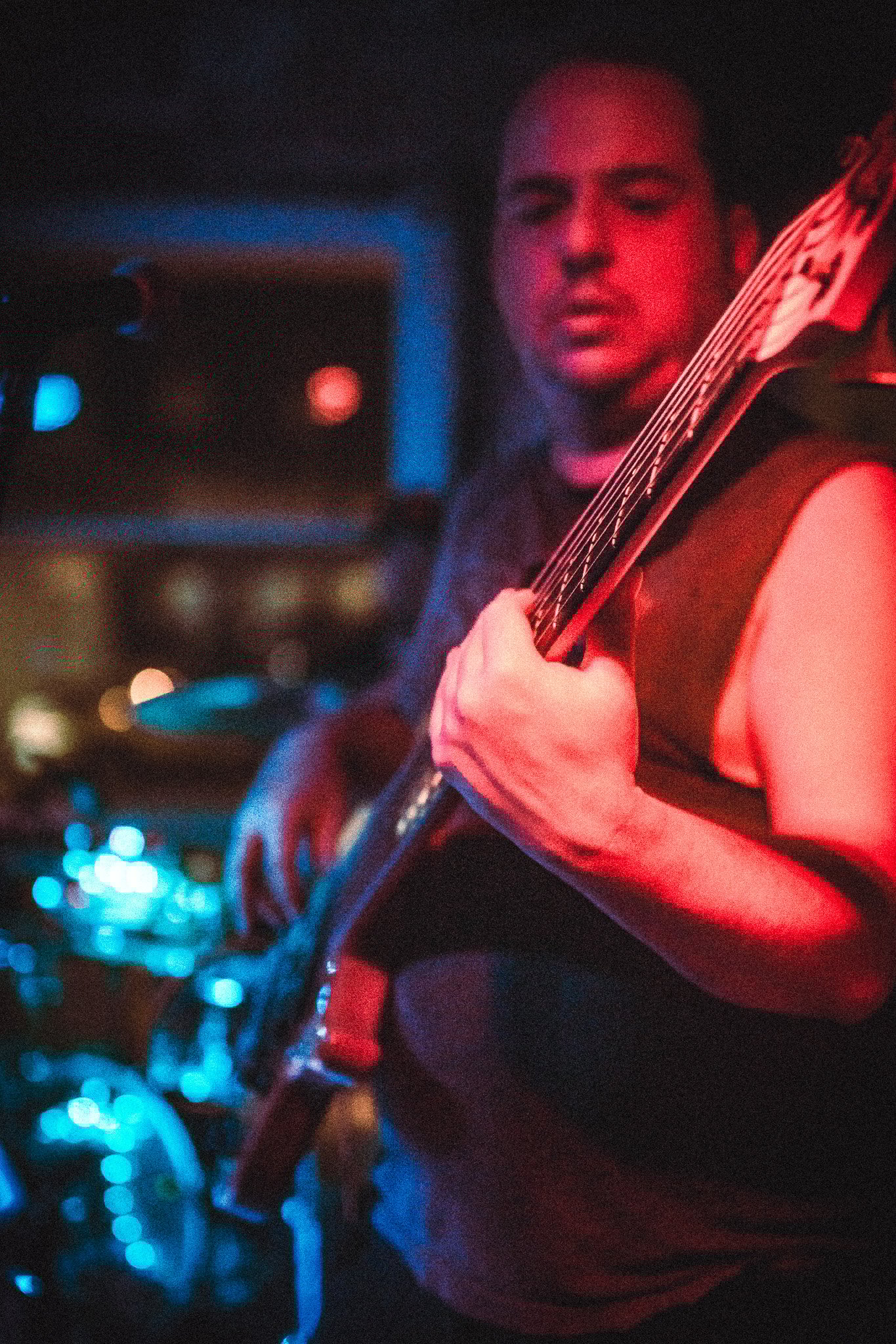 LUXE Man playing bass guitar under red stage lighting with blurred drum set in the background.