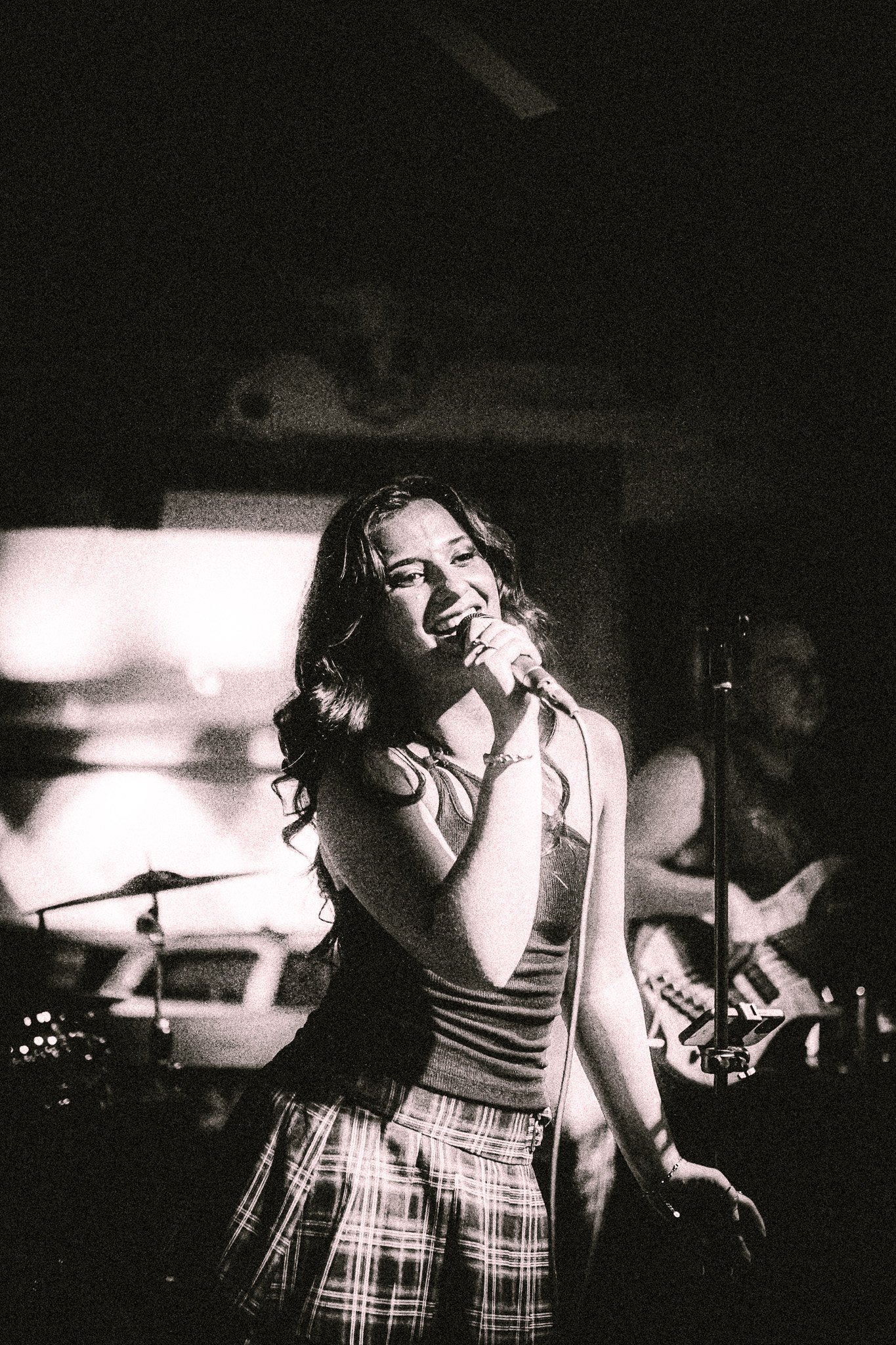 LUXE Woman singing passionately into a microphone on stage with a guitarist and drums in the background, in a black and white photo.