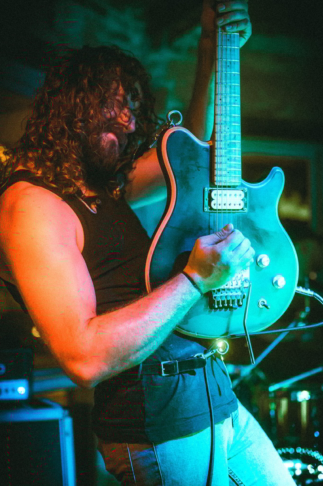 Man with long curly hair playing an electric guitar on stage with colorful lighting. LUXE 
