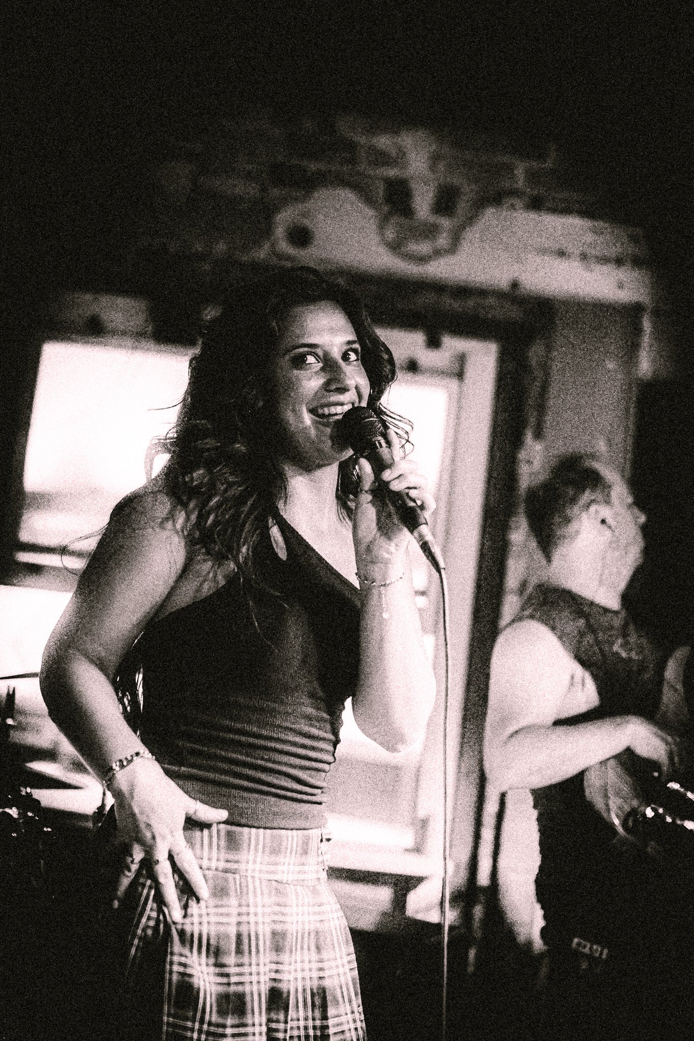 Smiling woman singing into a microphone on stage, wearing a sleeveless top and plaid skirt. LUXE 