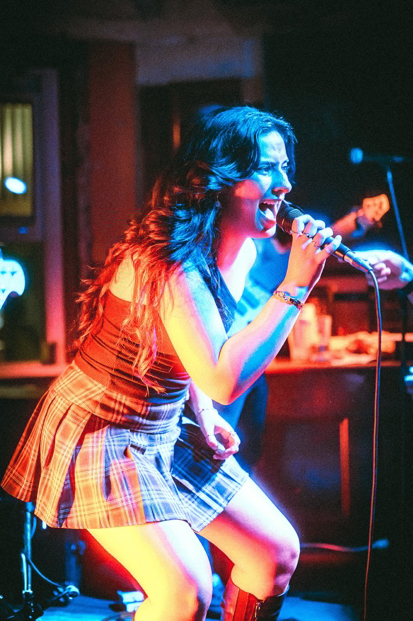 Female singer passionately performing on stage holding a microphone, wearing a plaid skirt and tank top under colorful stage lights. LUXE 