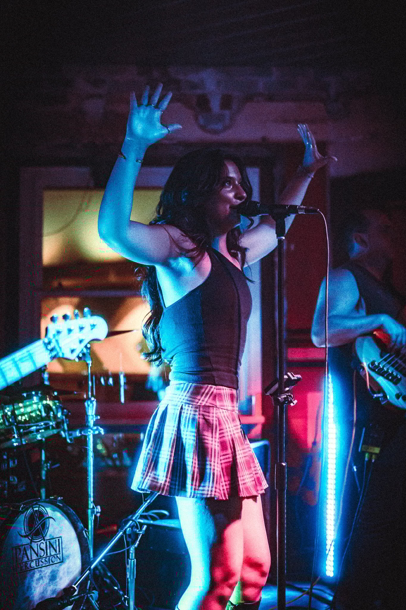 LUXE Female singer with long dark hair and plaid skirt performing on stage with hands raised, illuminated by blue and pink lighting.