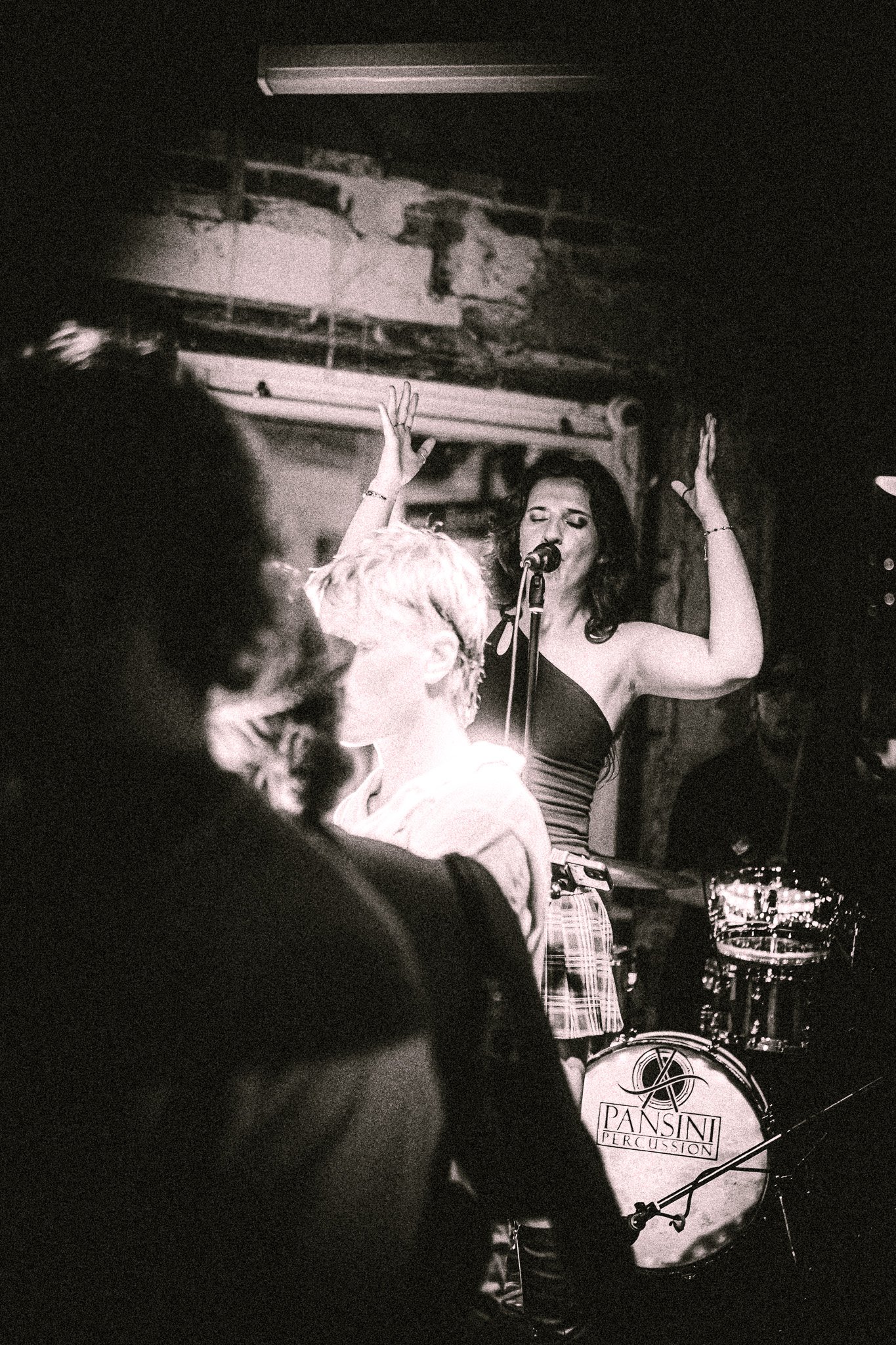 Black-and-white photo of a female singer with raised hands performing on stage in front of a drum set labeled 'Pansini Percussion.' LUXE 