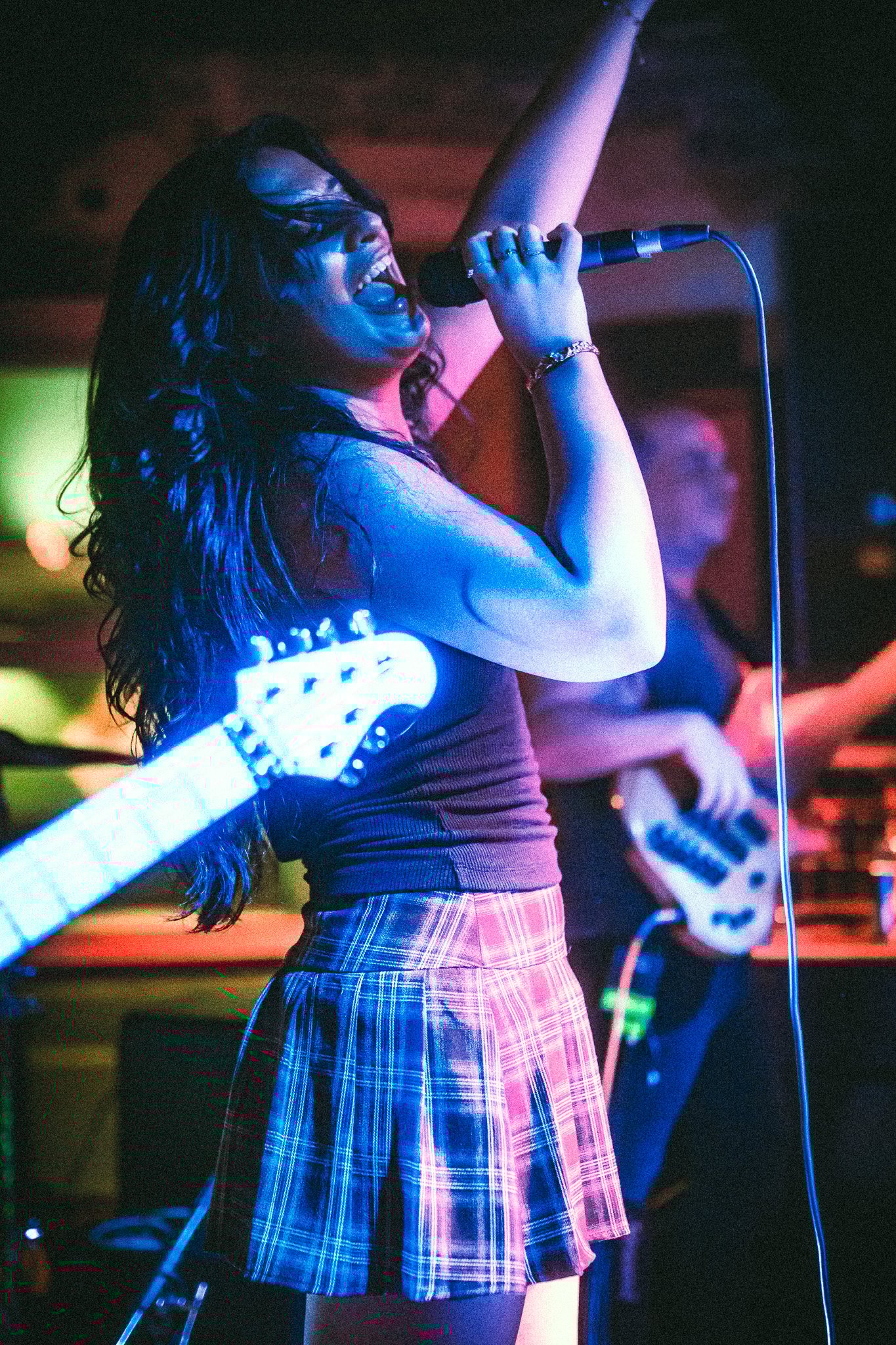 Female singer passionately performing with microphone on stage under blue lighting, with a guitarist in the background. LUXE 