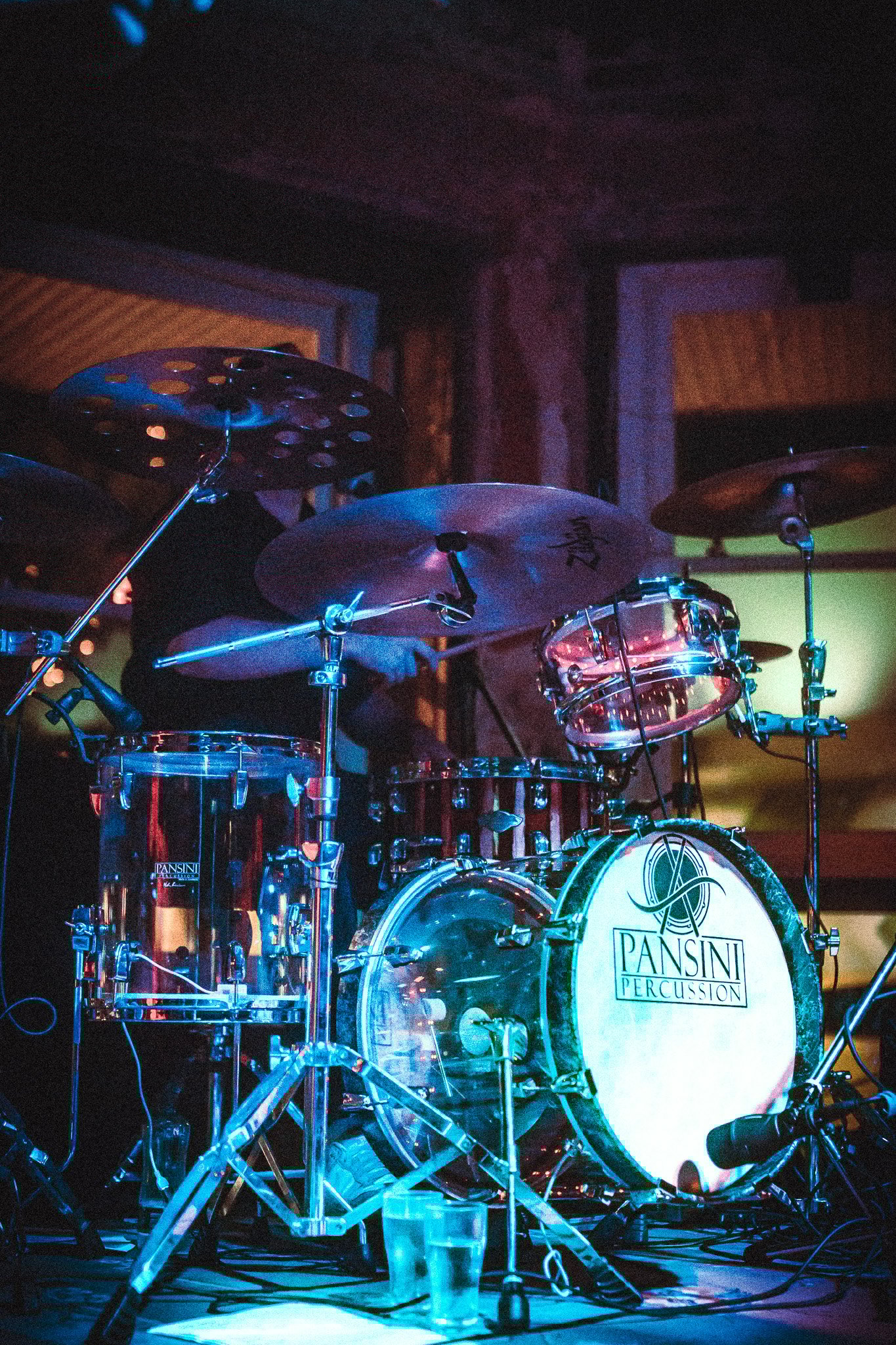 Close-up of a LUXE drum set with illuminated Pansini Percussion bass drum, cymbals, and microphones in a dimly lit room.