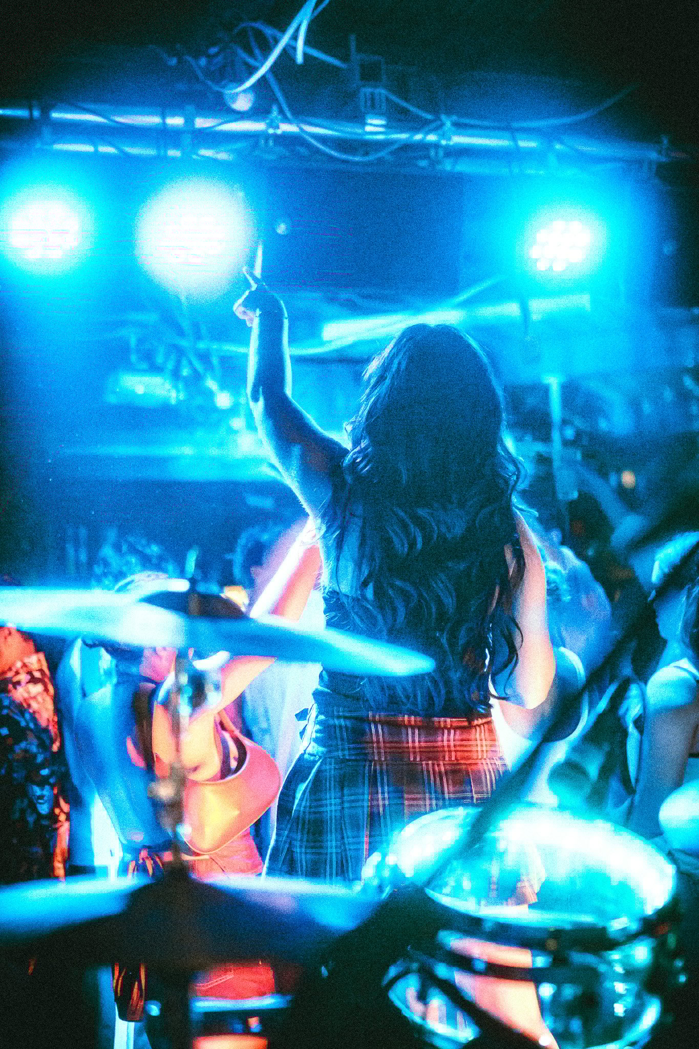 LUXE Person with long hair and plaid skirt raising one arm in a blue-lit concert or party crowd viewed from behind with drum set in foreground.