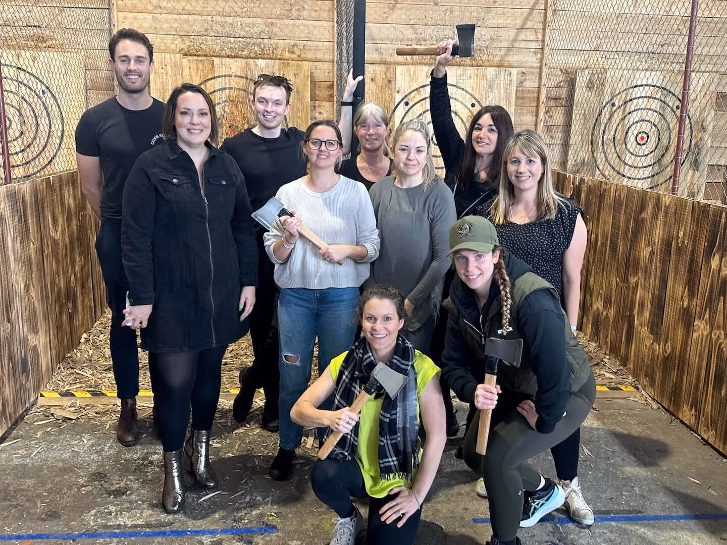 Group of ten people at an indoor axe throwing venue, some holding axes and standing in front of wooden targets.