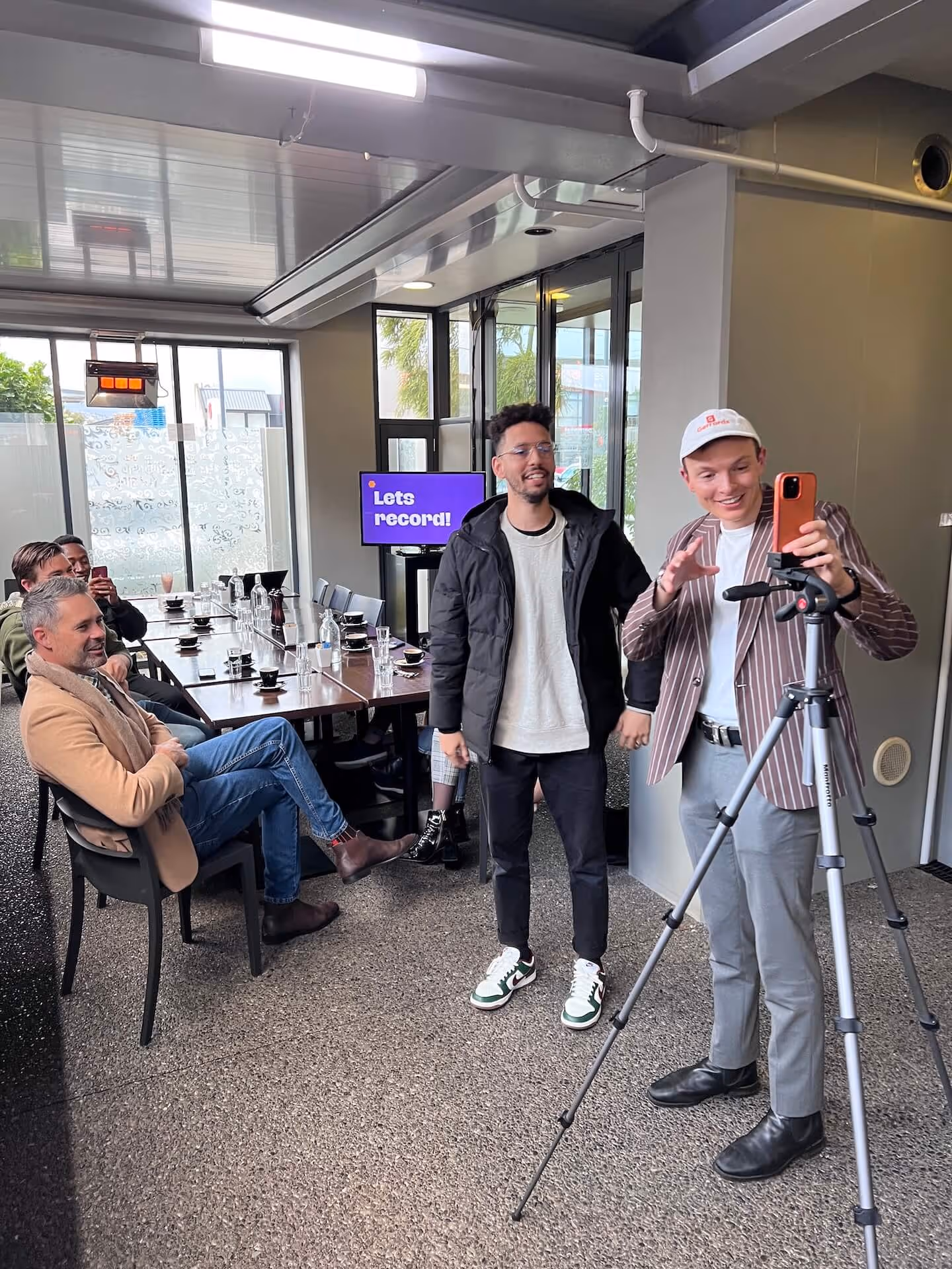 A group of men indoors with one man setting up a smartphone on a tripod to record, and others sitting around a table with a screen showing 'Lets record!' in the background.