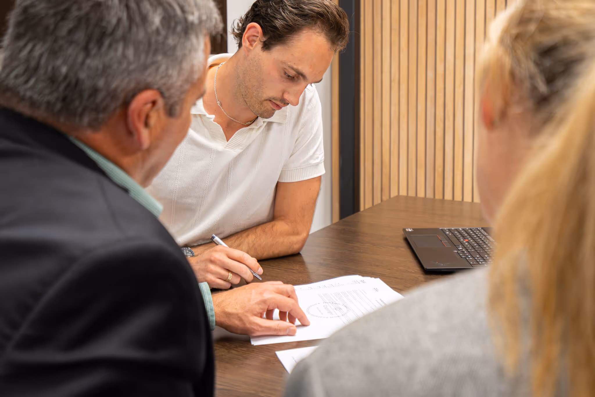 A man in a white shirt reviews and signs documents at a table while two other people watch.