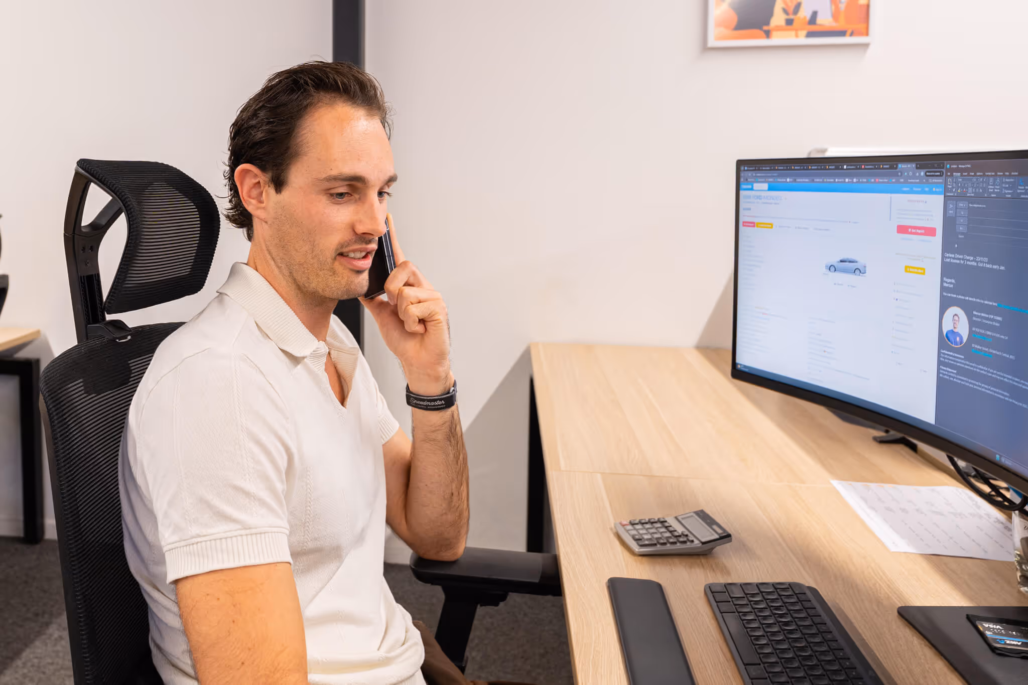 Man in a white polo shirt sitting at a desk talking on a smartphone with a curved monitor displaying a car report and email on the screen.