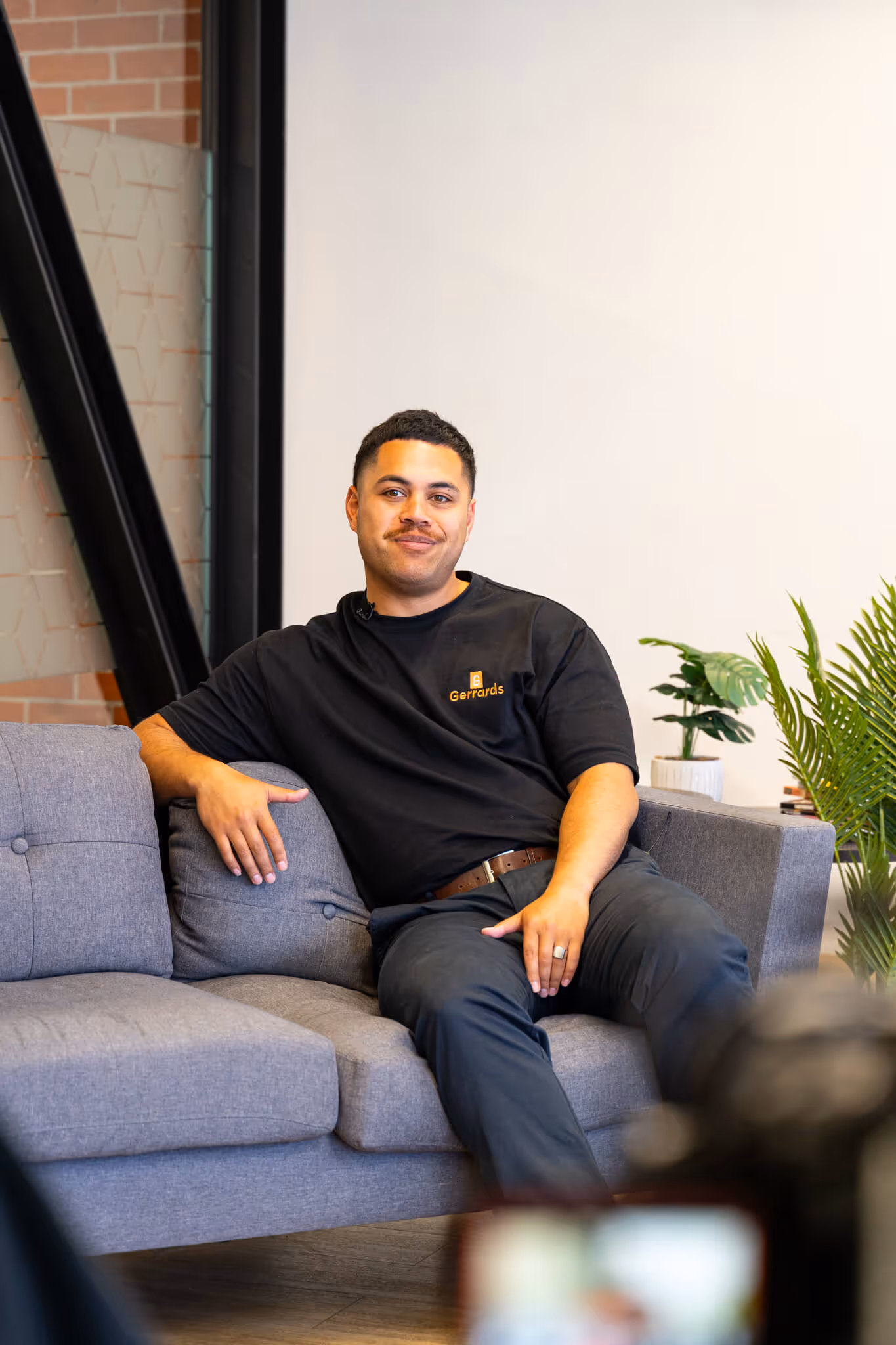 Young man with a mustache wearing a black Gerrards t-shirt sitting relaxed on a gray couch in a modern room with plants.