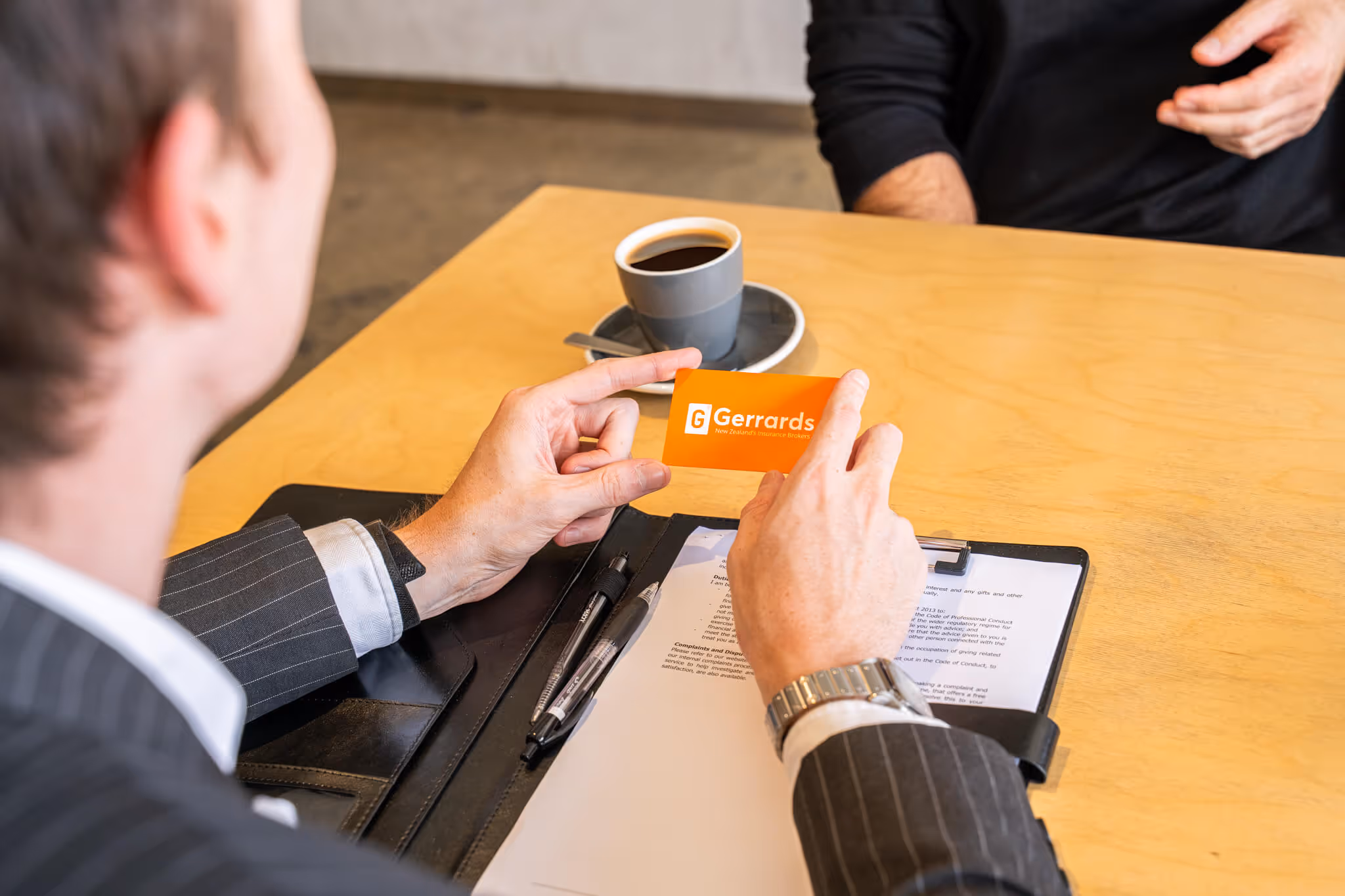 Person in a pinstripe suit holding an orange Gerrards business card over a table with a clipboard of documents and a cup of coffee.