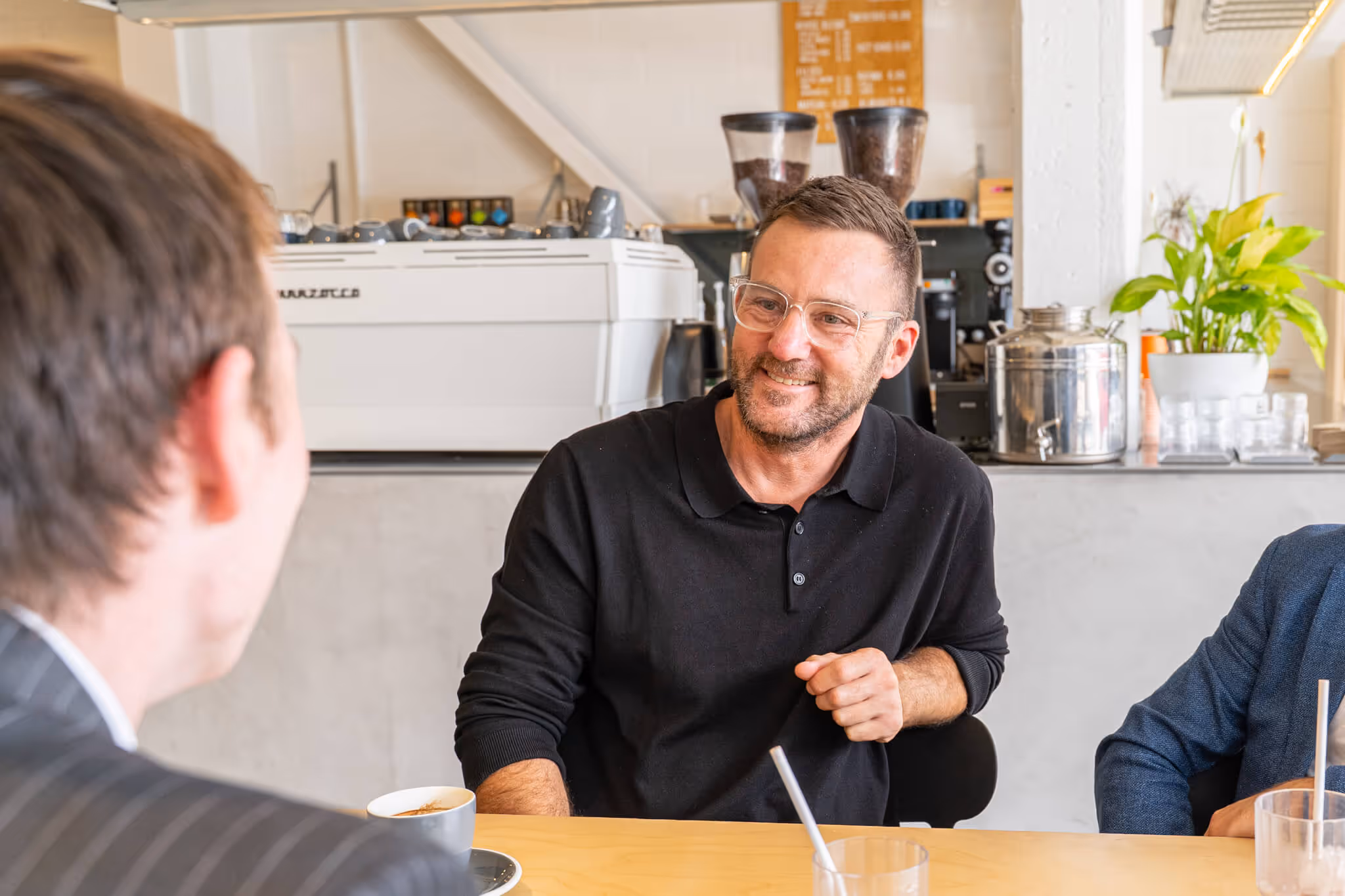 Smiling man wearing glasses and a black shirt talking to another man in a café.