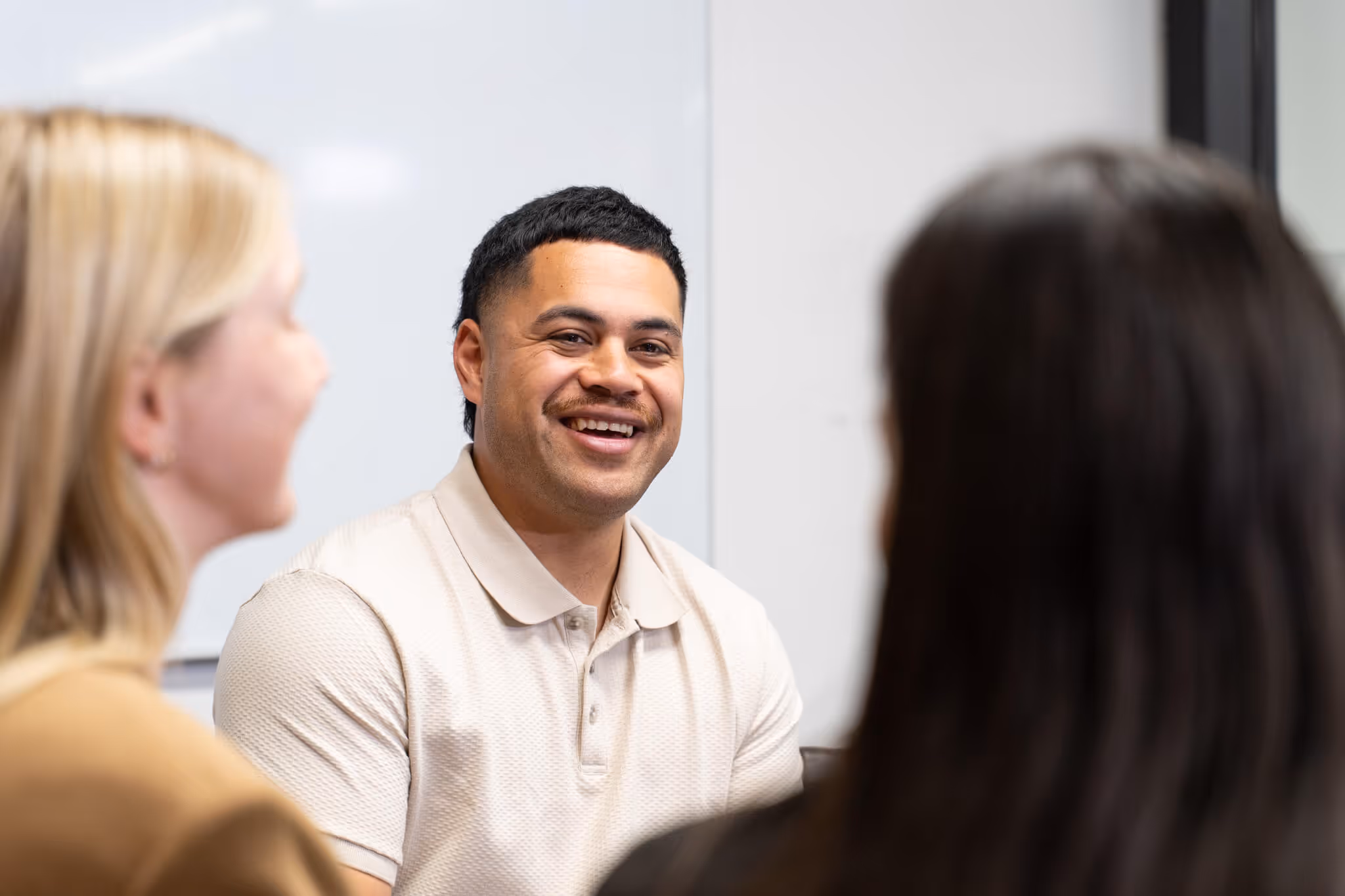 Smiling man in a white polo shirt engaged in conversation with two women in an office setting.