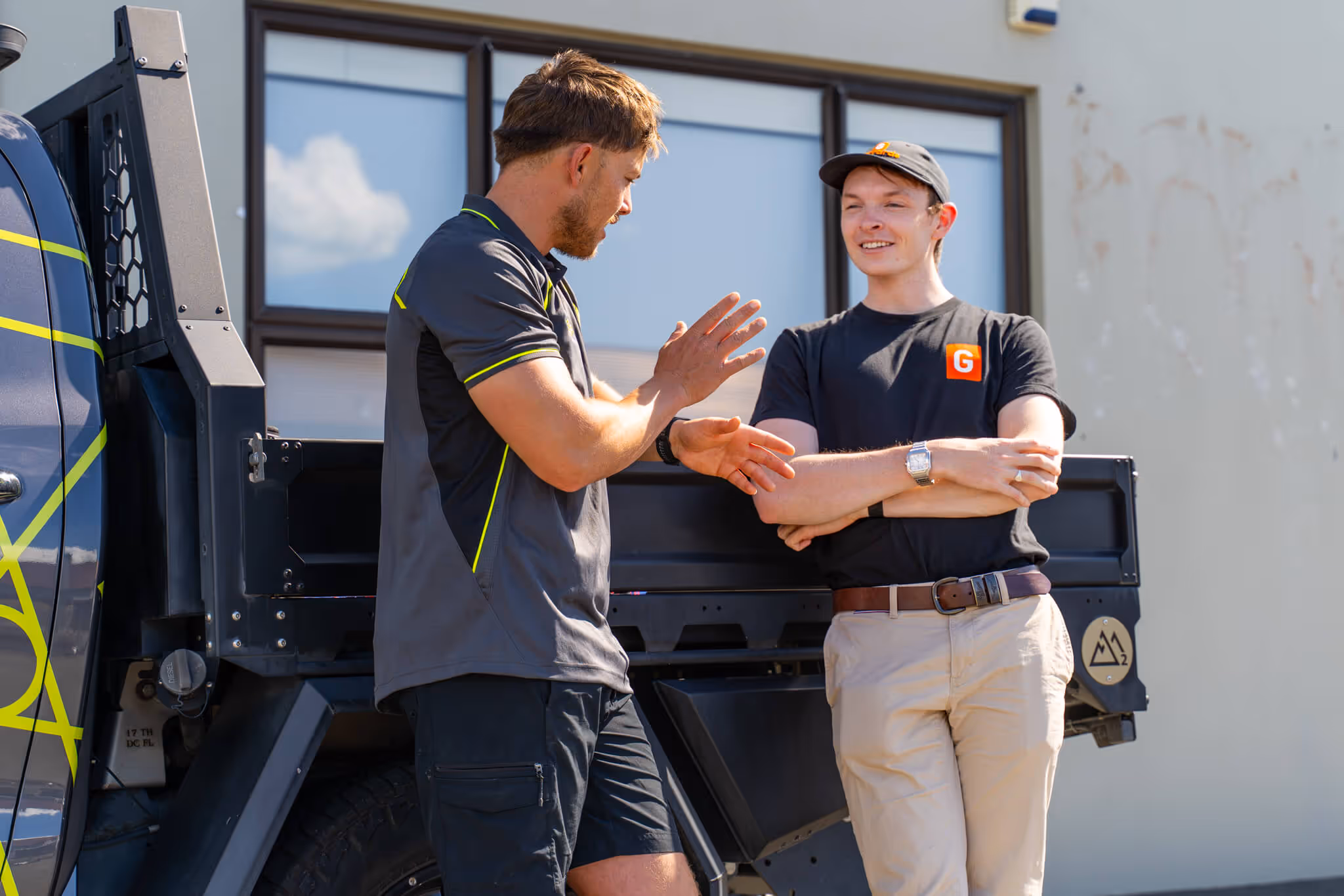 Two men in casual workwear standing and chatting by a truck on a sunny day.