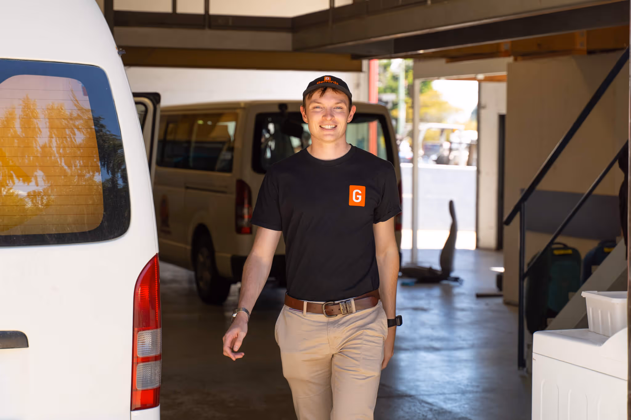 Smiling young man wearing a black Gerrards cap and t-shirt walking indoors near white vans.