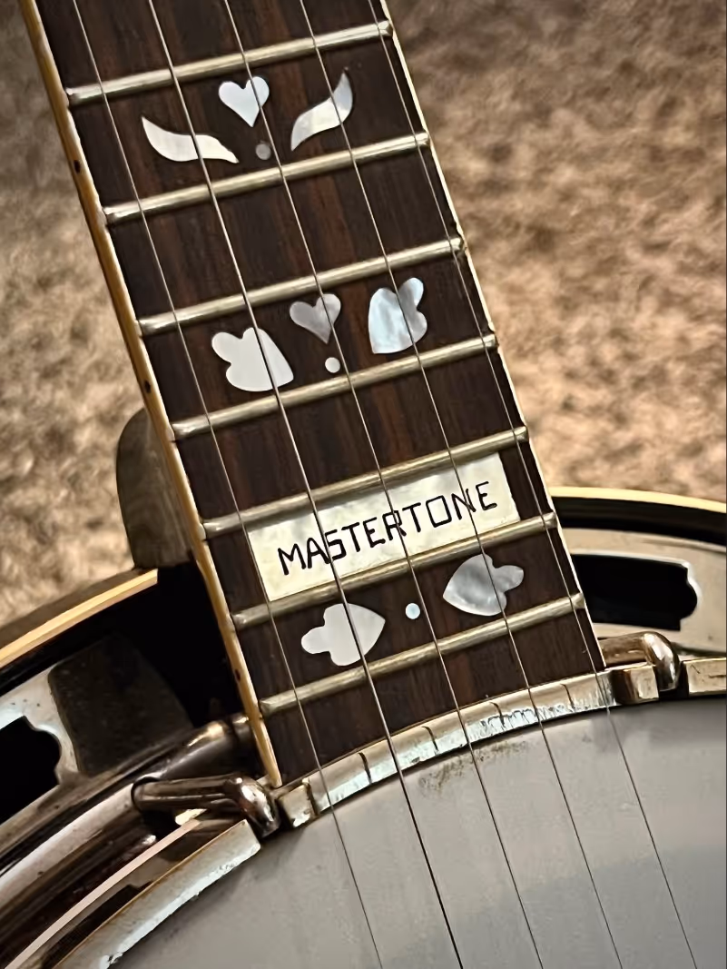 Close-up of a banjo fretboard with decorative inlays and the word 'MASTERTONE' on a mother-of-pearl plate.