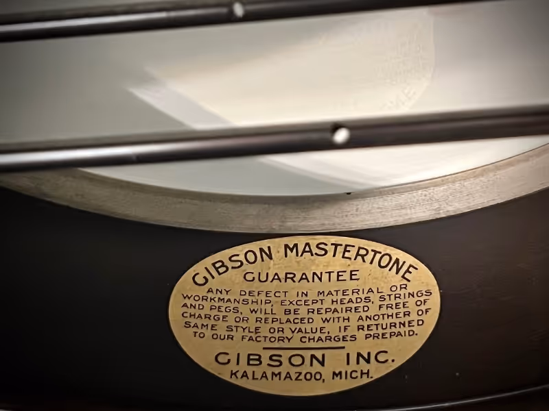 Close-up of oval Gibson Mastertone guarantee label inside a banjo rim with two metal coordinator rods above it.
