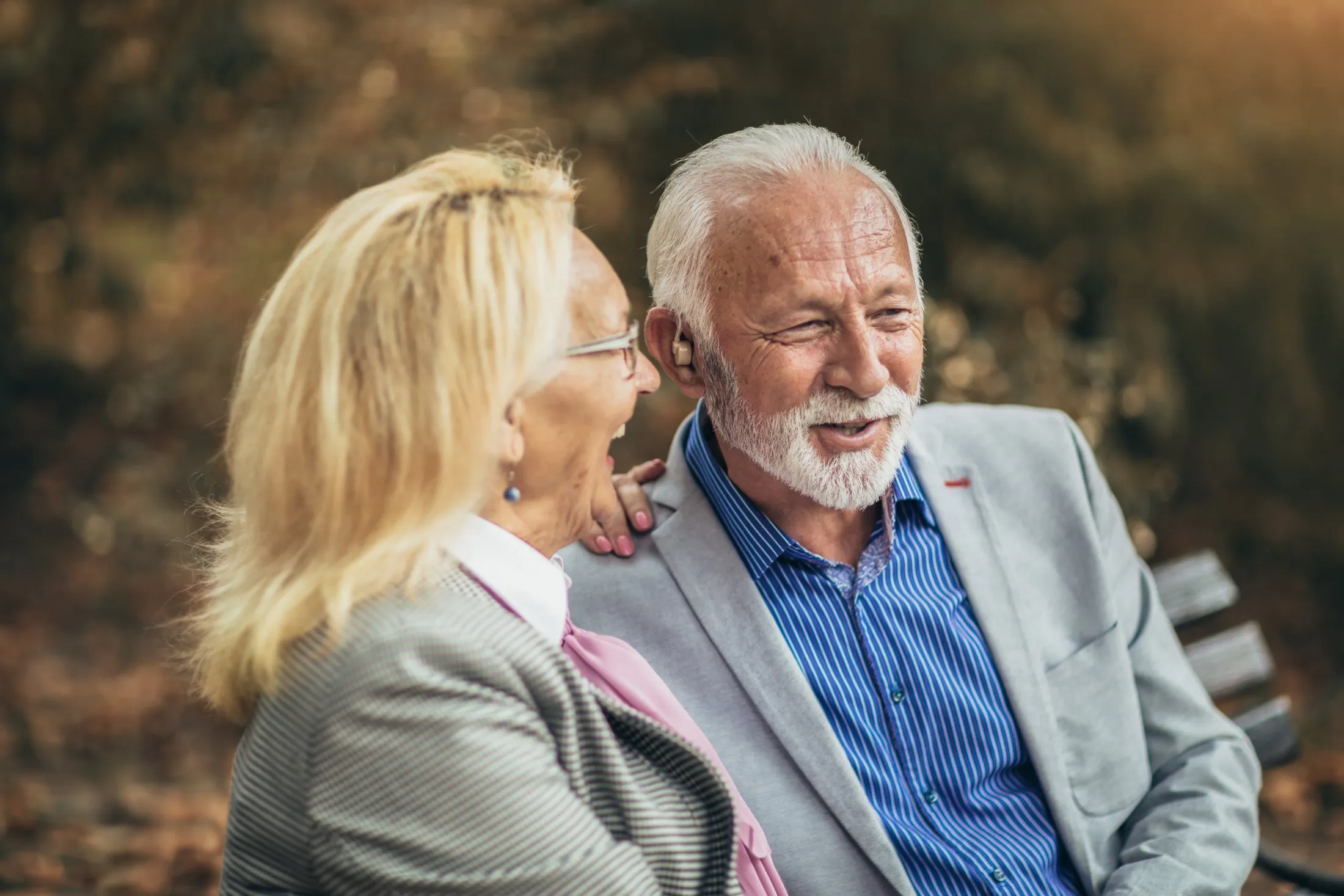 Elderly man and woman sitting on a bench outdoors, smiling and having a conversation.