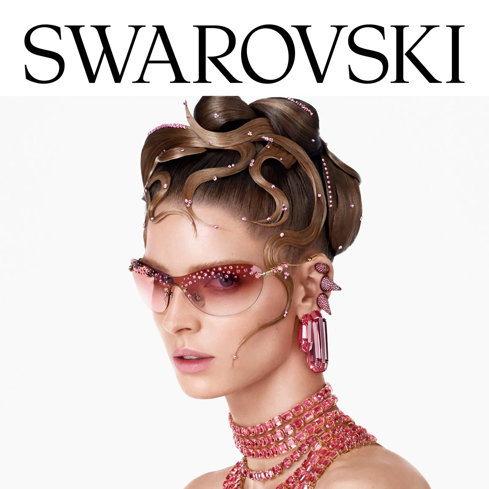Woman with sculpted hair adorned with pink crystals, wearing pink-tinted sunglasses and matching pink crystal jewelry including earrings and a multi-layered necklace, under Swarovski logo.